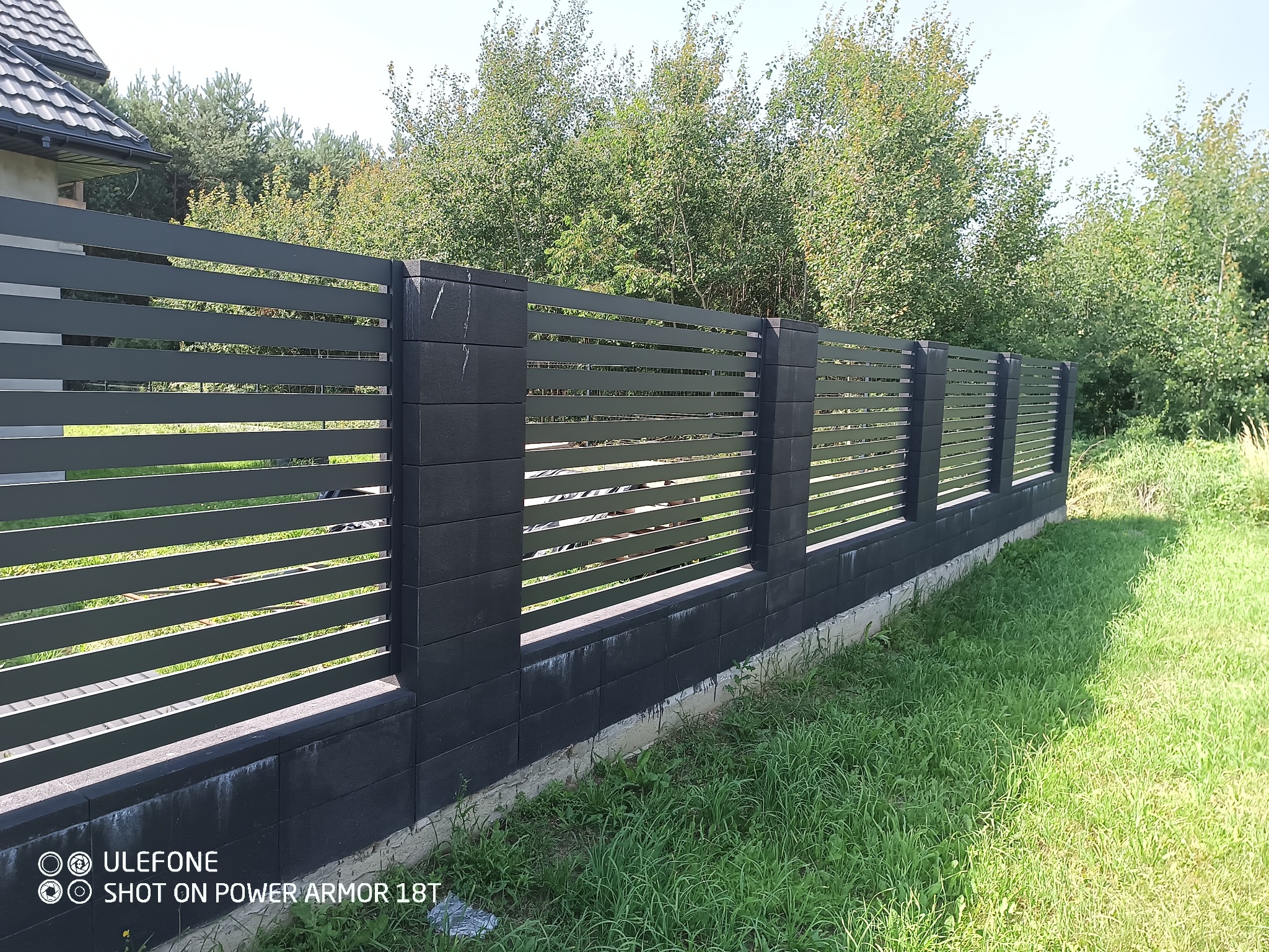 Nowoczesne ogrodzenie panelowe w kolorze grafitowym z betonowymi słupkami, otoczone zielenią. Widok na fragment domu i drzewa w tle.