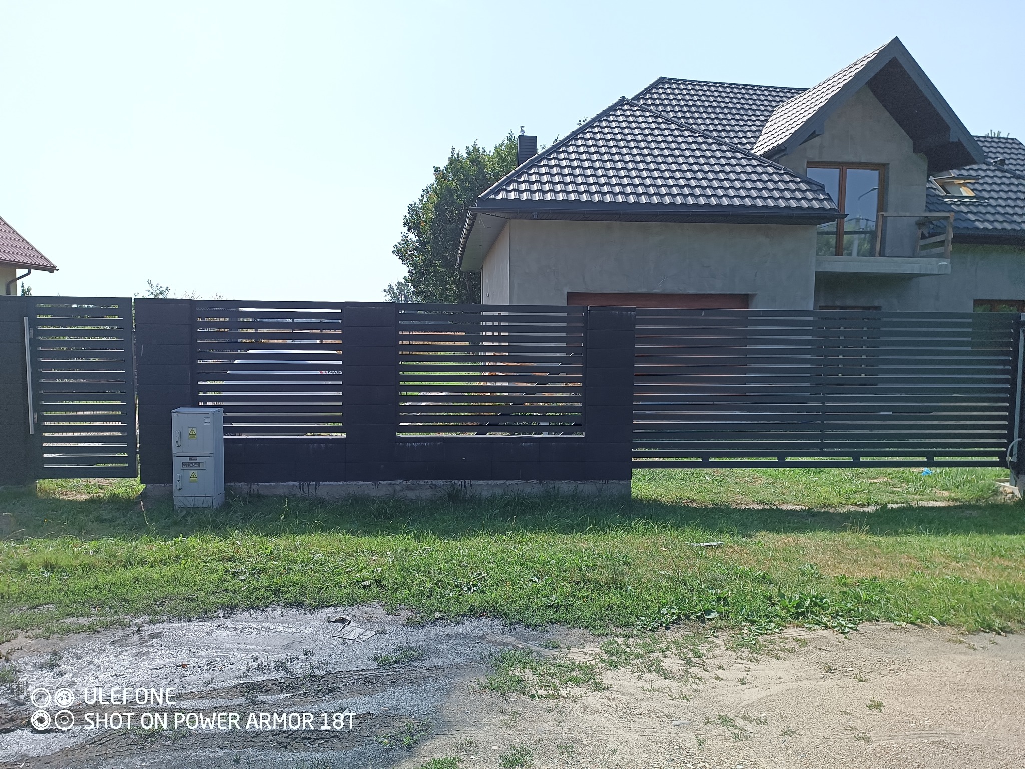 Nowoczesne ogrodzenie posesji z poziomych, ciemnych paneli, z widoczną skrzynką elektryczną i fragmentem domu w tle, w słoneczny dzień.