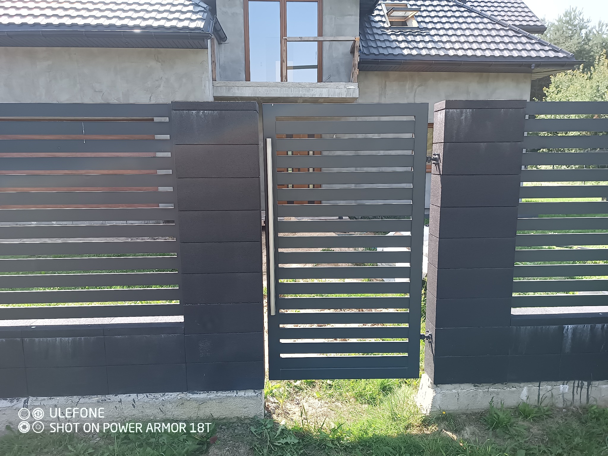 Nowoczesna, szara brama panelowa z poziomymi szczeblami, częściowo otwarta. W tle dom w stanie surowym i fragment ogrodzenia z elementami drewnianymi.