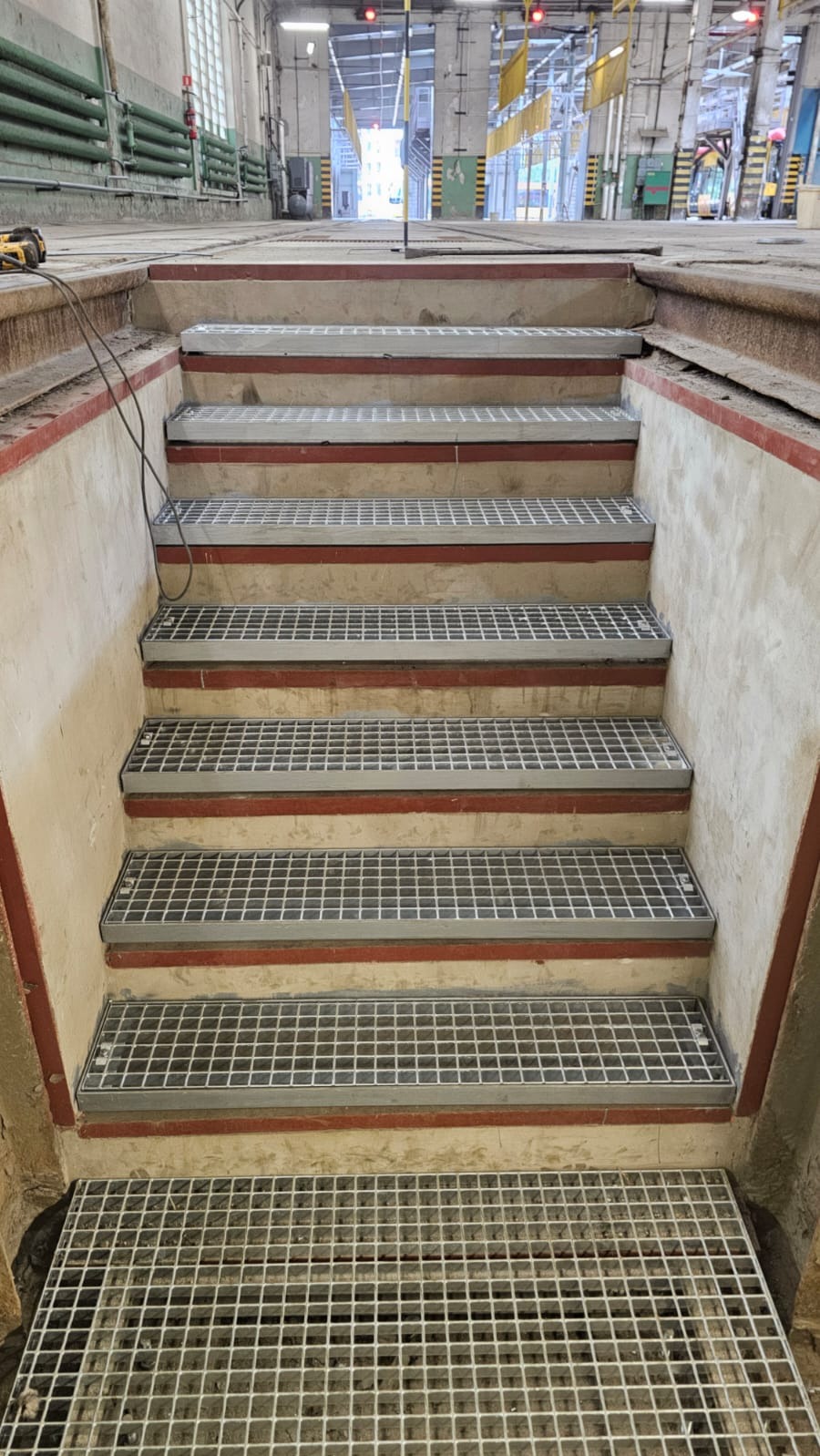 Metalowe schody kratowe w surowym wnętrzu hali przemysłowej, widok z dołu. Czerwone akcenty na stopniach, w tle widoczne światło dzienne.