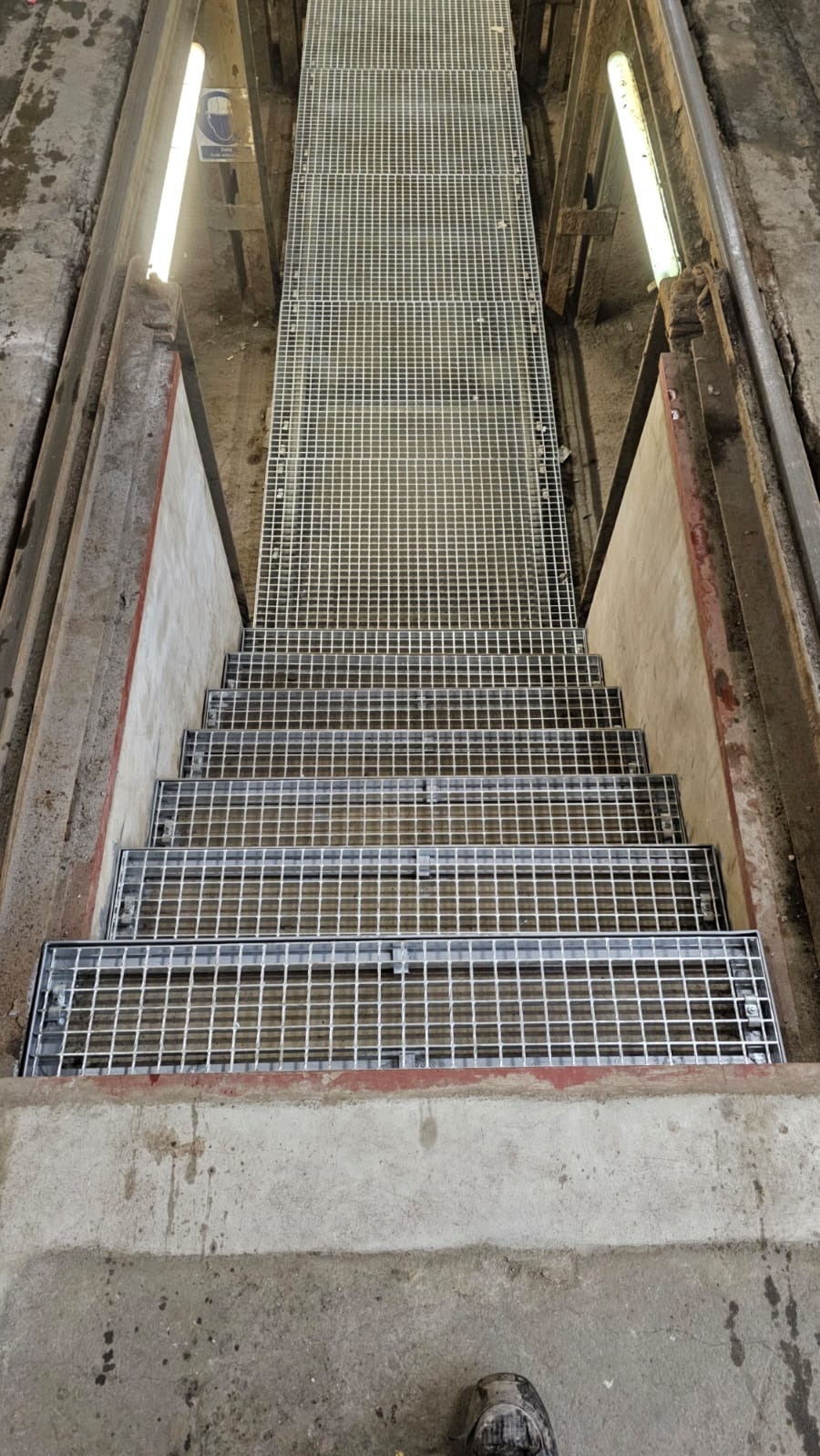 Metalowe schody kratowe w surowym otoczeniu przemysłowym, widziane z góry. Widoczne oświetlenie fluorescencyjne i fragment buta w dolnej części kadru.