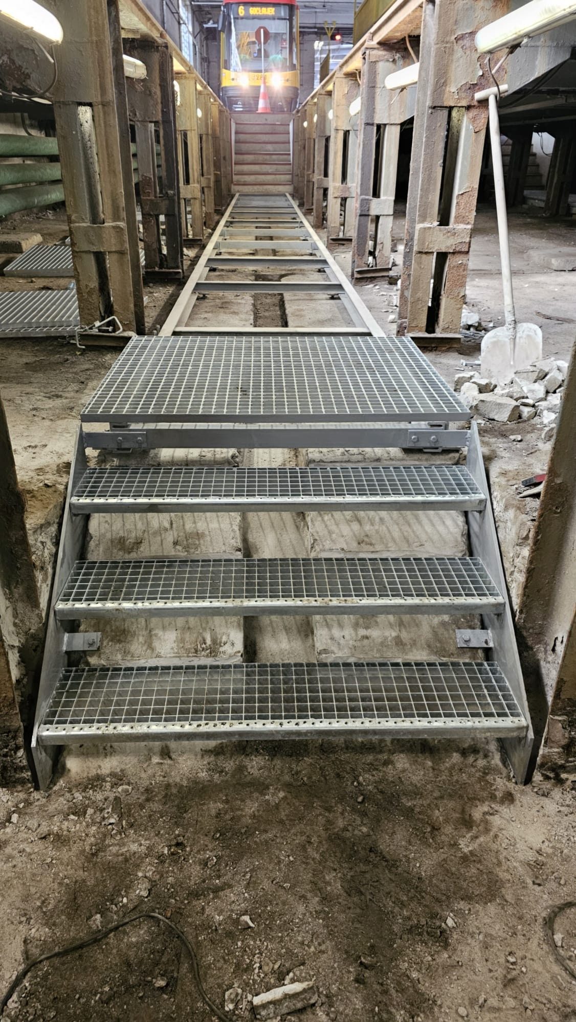 Metalowe schody kratowe prowadzące w dół, widok z dołu z perspektywą na tramwaj w tle. Konstrukcja stalowa w surowym, industrialnym otoczeniu.