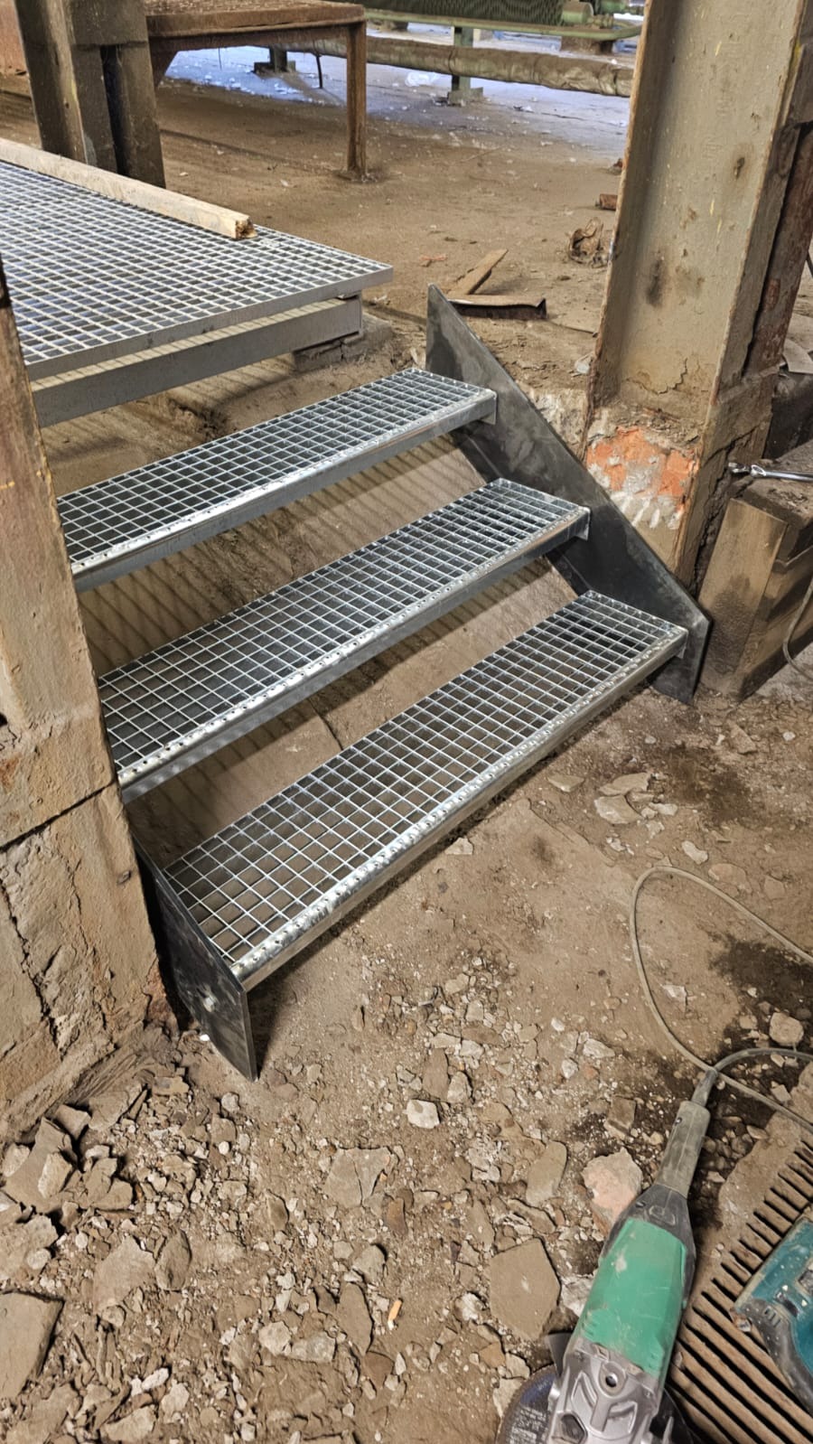 Metalowe schody kratowe w surowym wnętrzu. Widoczne narzędzia pracy na podłodze. Detal konstrukcji schodów z metalową siatką, w trakcie montażu lub po nim.