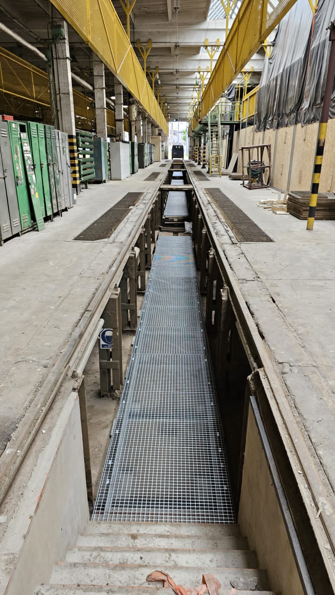 Długa, metalowa krata w kanale warsztatowym z widocznymi schodami wejściowymi, żółtymi suwnicami i rzędami szafek w tle hali.