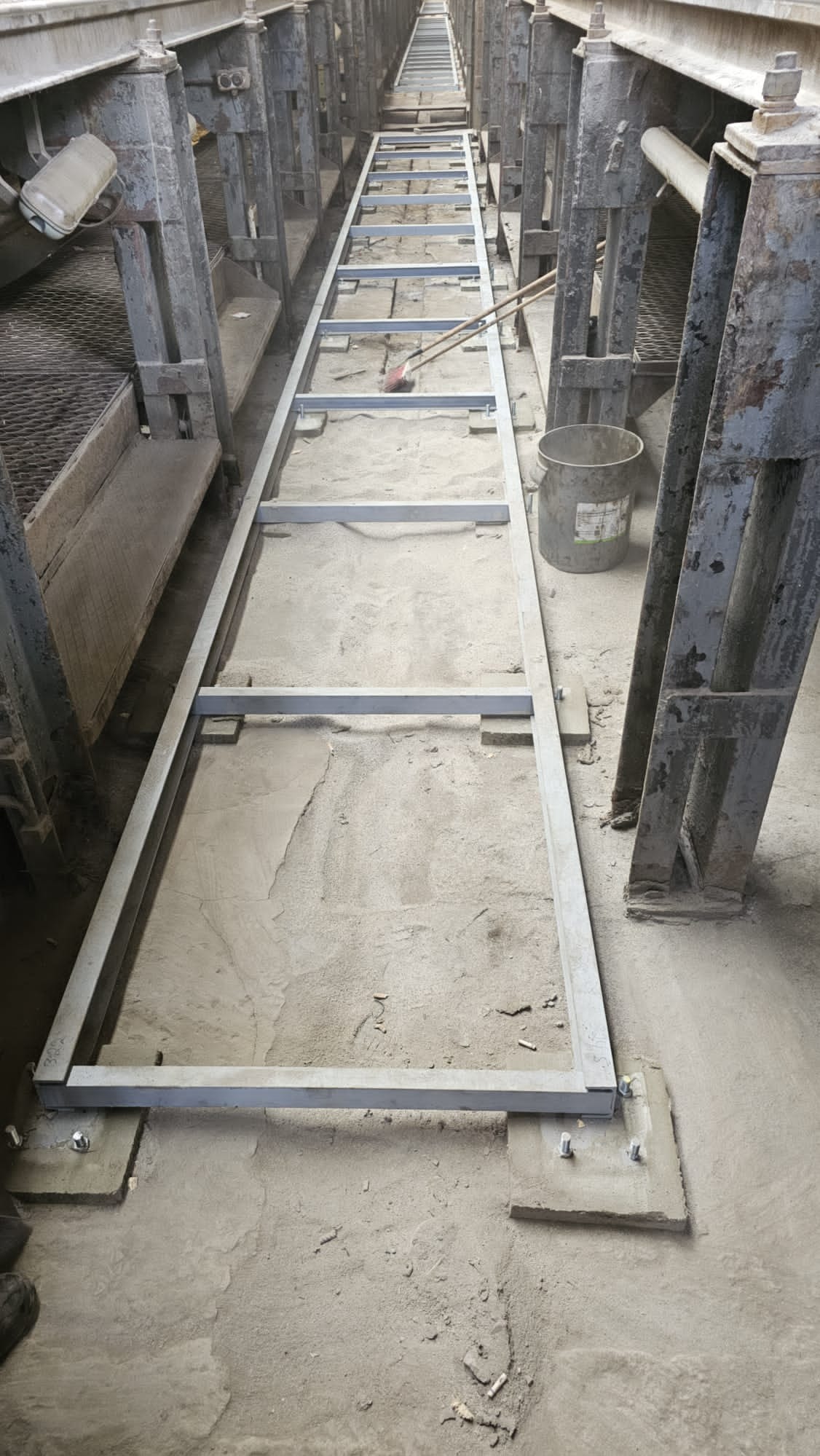 Długa, stalowa rama konstrukcji w trakcie montażu, przytwierdzona śrubami do betonowych fundamentów. Widoczne elementy konstrukcji hali.