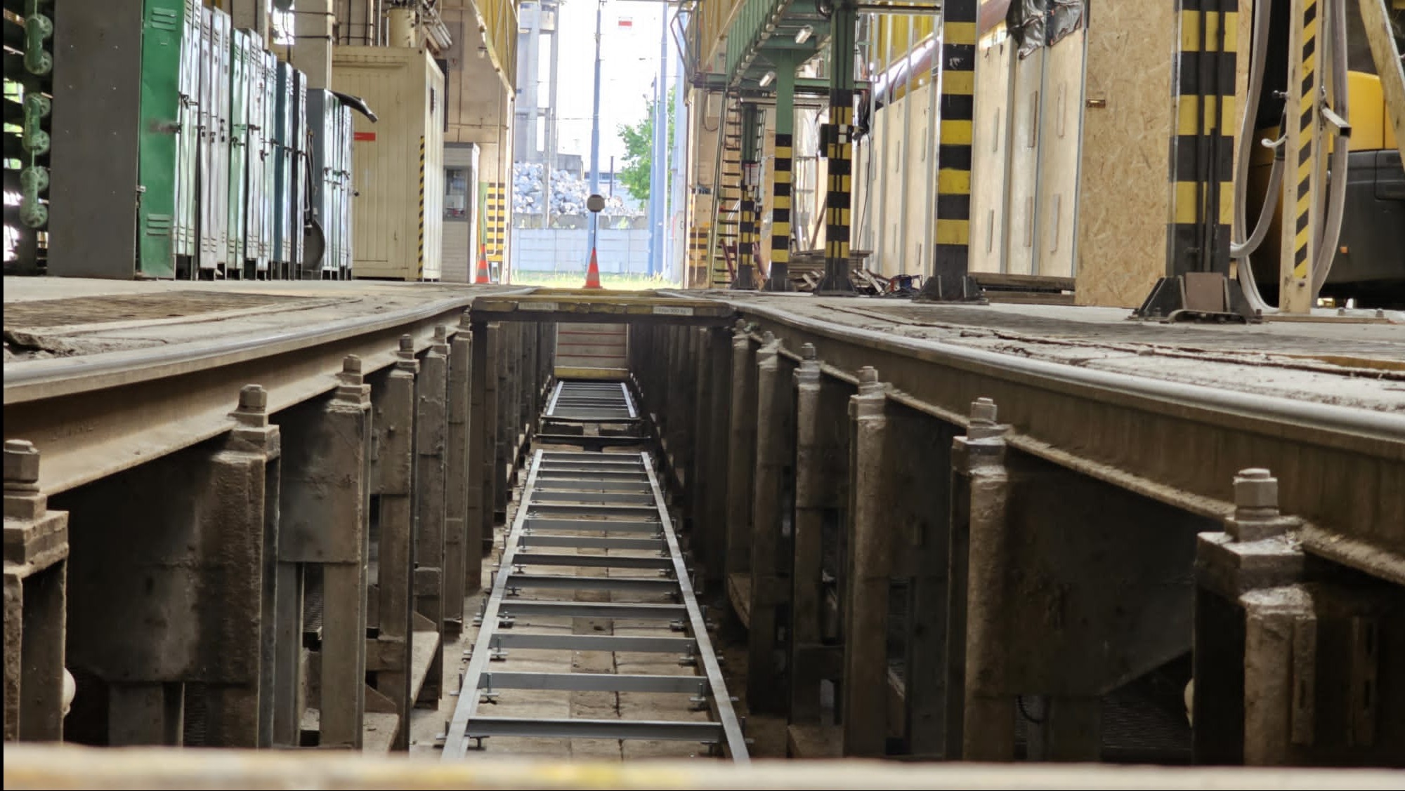 Widok na metalową konstrukcję w hali, z perspektywy zagłębienia w podłodze. Widoczne szyny, metalowe podpory i drabina w głębi. Oświetlenie dzienne.