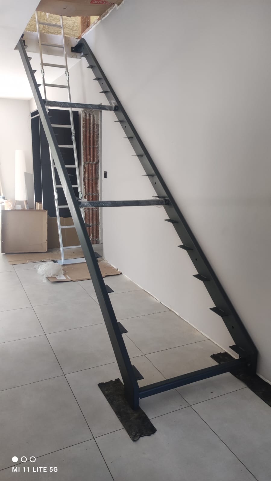 Surowe, czarne schody metalowe ze stopniami w trakcie montażu w nowym budynku, z widoczną drabiną prowadzącą na strych i elementami instalacji elektrycznej.