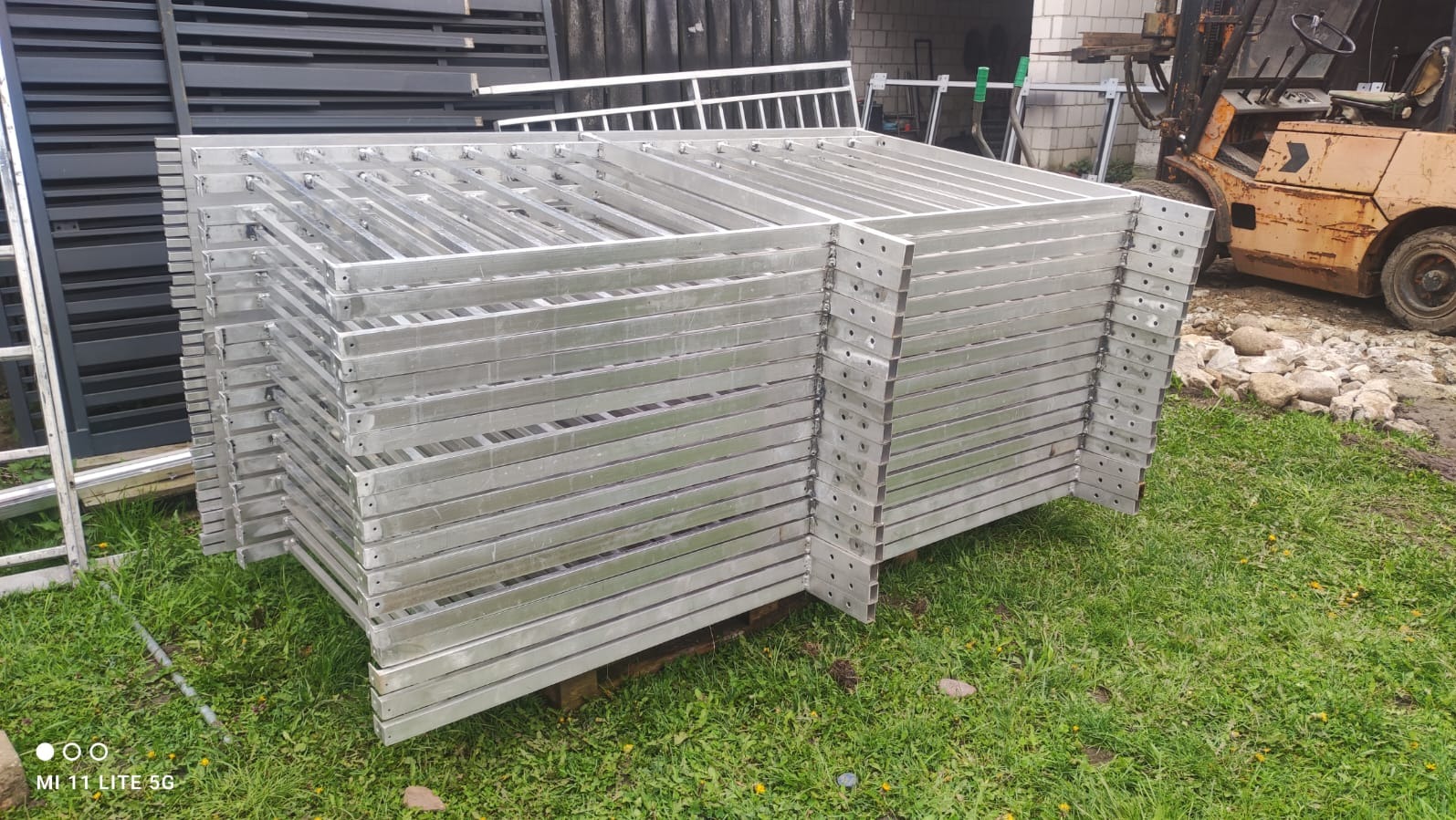 Stos srebrnych, metalowych ram ogrodzeniowych z otworami, ułożonych na palecie na trawie, z wózkiem widłowym w tle. Elementy gotowe do montażu.