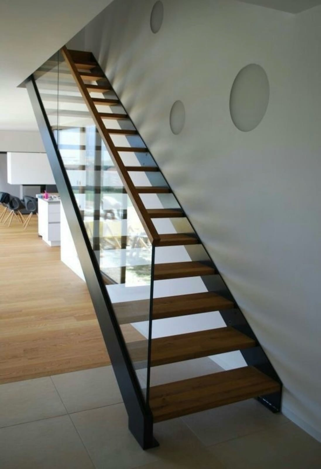 Nowoczesne schody z drewnianymi stopniami i szklaną balustradą, czarna konstrukcja, minimalistyczny design, widok z dołu, jasne wnętrze z parkietem.