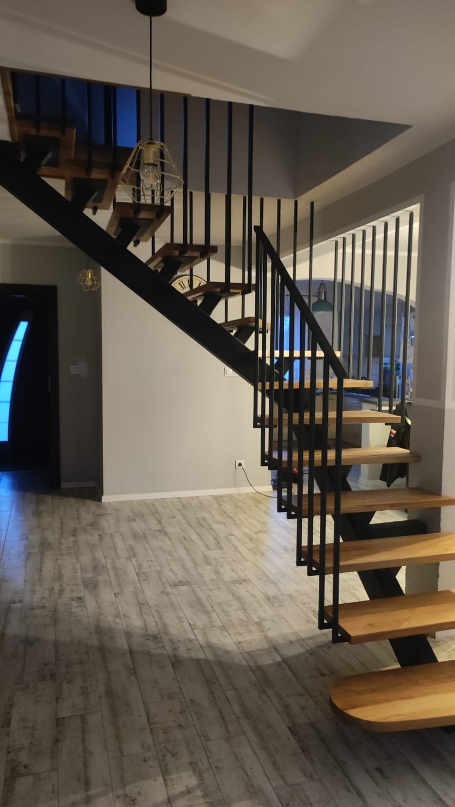 Nowoczesne schody metalowe z drewnianymi stopniami, czarne balustrady pionowe, widok z dołu. Oświetlenie punktowe. Podłoga imitująca drewno.