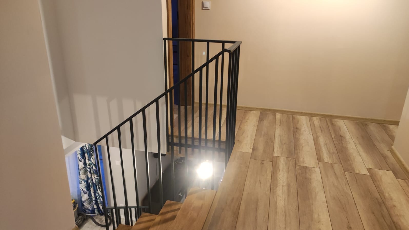 Nowoczesna, czarna poręcz schodowa z pionowymi szczebelkami, zamontowana w jasnym wnętrzu z drewnopodobną podłogą. Widok z góry na schody i fragment ściany.