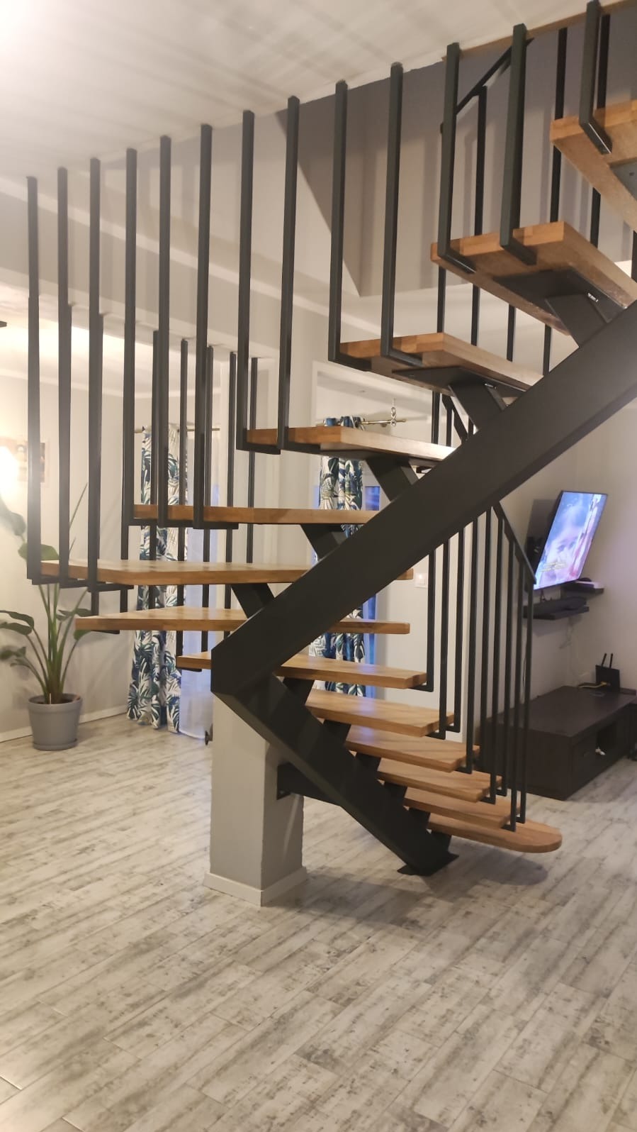 Nowoczesne schody metalowe z drewnianymi stopniami, czarna konstrukcja, minimalistyczny design. Widok z dołu, jasne wnętrze z rośliną w tle.