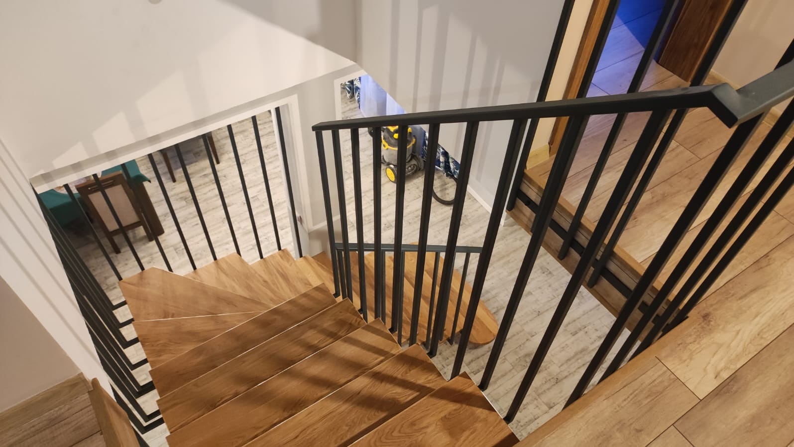 Drewnopodobne schody z czarną, metalową balustradą, widziane z góry. W tle widoczny odkurzacz i fragmenty innych pomieszczeń.