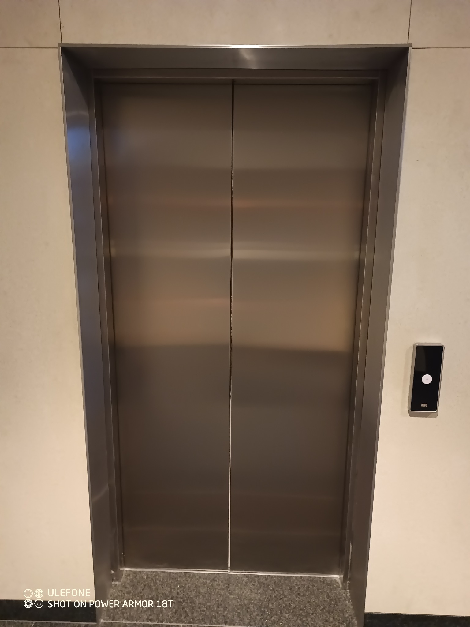 Metalowe, dwuskrzydłowe drzwi windy w srebrnej ramie, z panelem sterowania po prawej stronie, na tle jasnych ścian i szarej podłogi.