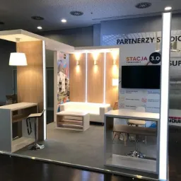 Aranżacja stoiska targowego z drewnianymi panelami, białymi meblami i podświetleniem LED, prezentująca ofertę osiedla mieszkaniowego na tle grafiki z partnerami i wizualizacją stadionu.