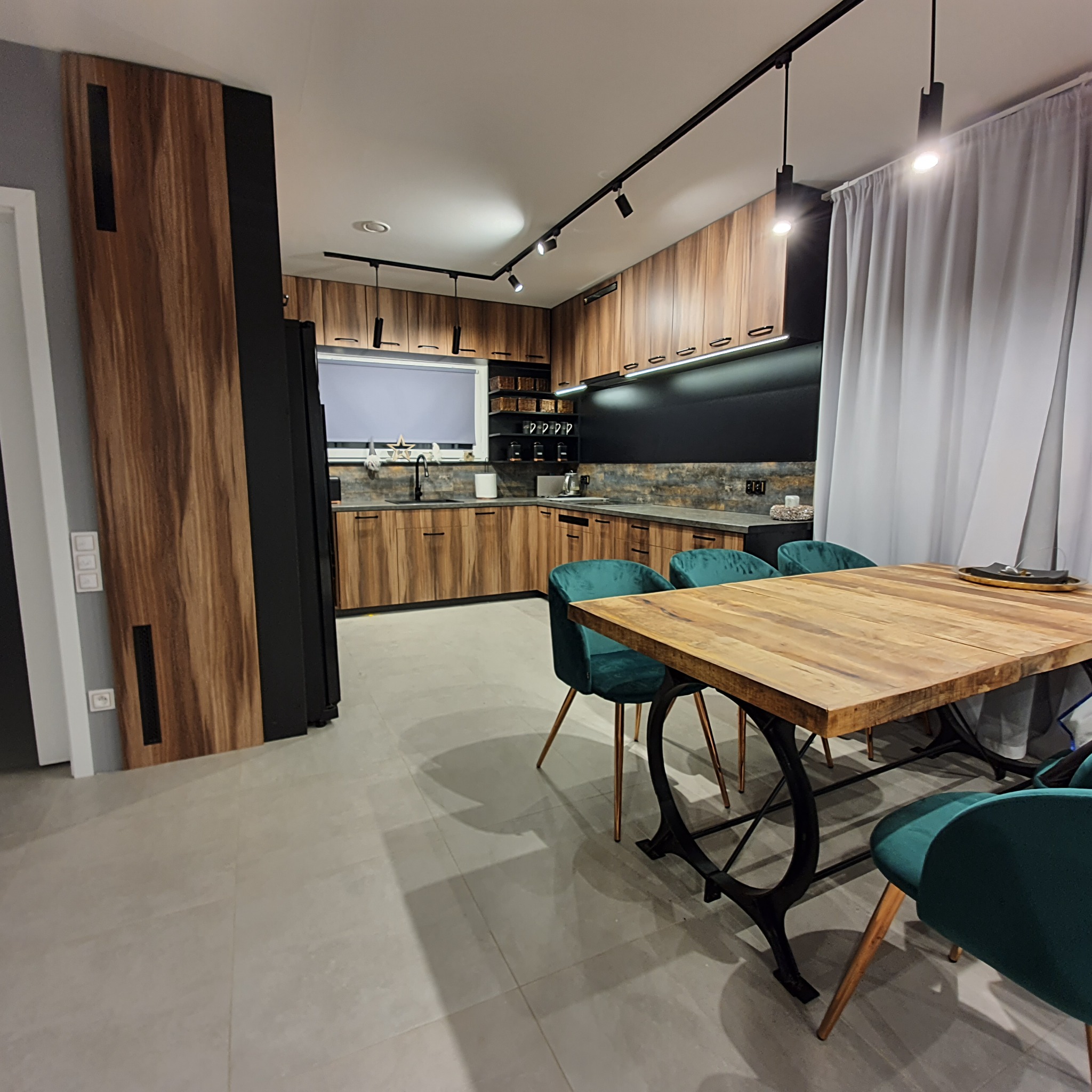 Nowoczesna kuchnia z drewnianymi frontami i czarnymi akcentami w Łodzi, oświetlenie szynowe, stół z drewnianym blatem i zielonymi krzesłami.