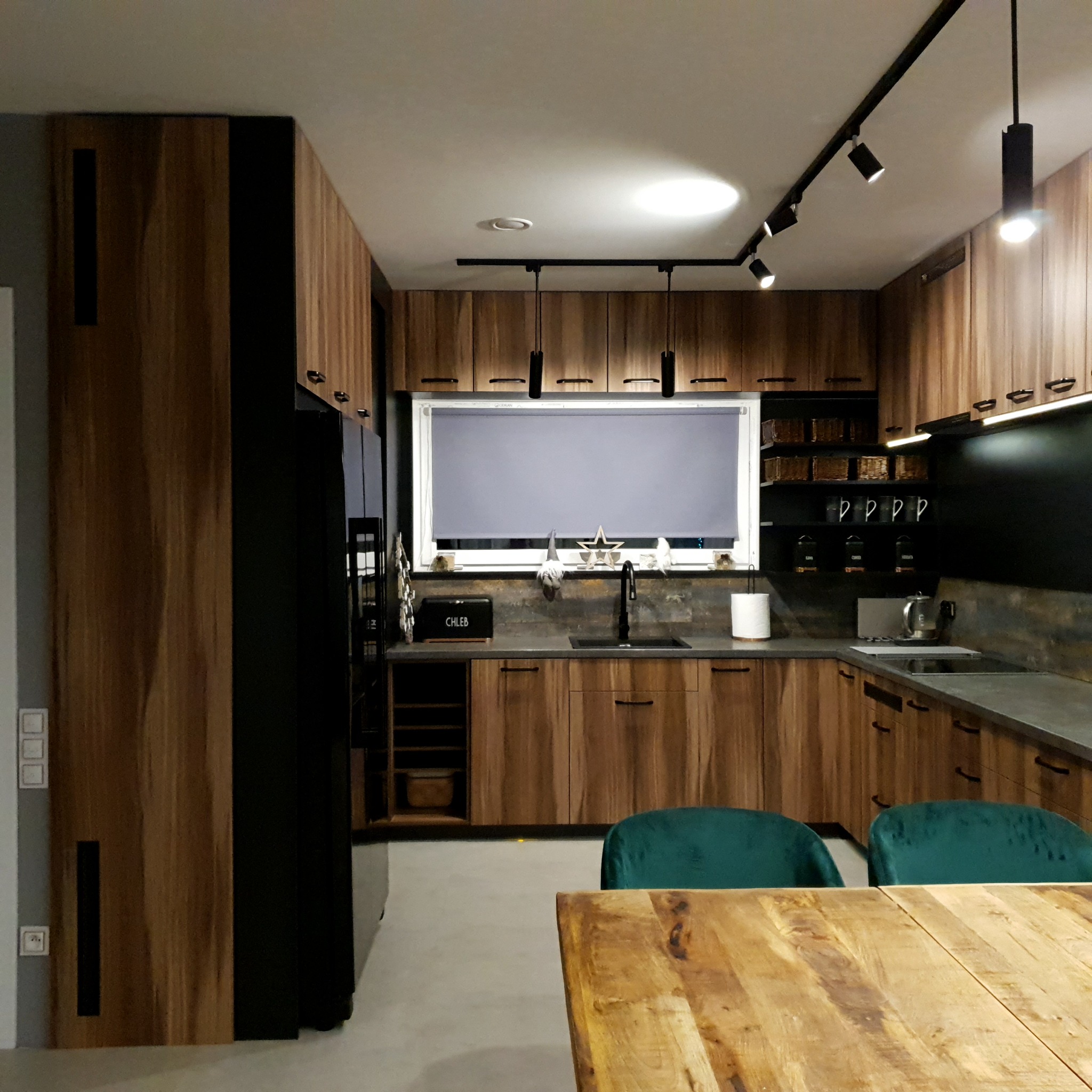 Nowoczesna kuchnia z drewnianymi frontami szafek, czarnymi akcentami i oświetleniem punktowym, widoczne zielone krzesła i drewniany stół na pierwszym planie.