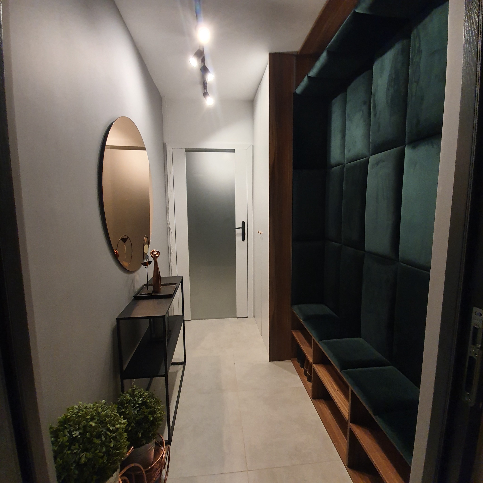 Nowoczesny przedpokój z owalnym lustrem, czarnym stolikiem z dekoracjami i zabudowaną wnęką z siedziskiem obitym ciemnozielonym welurem.