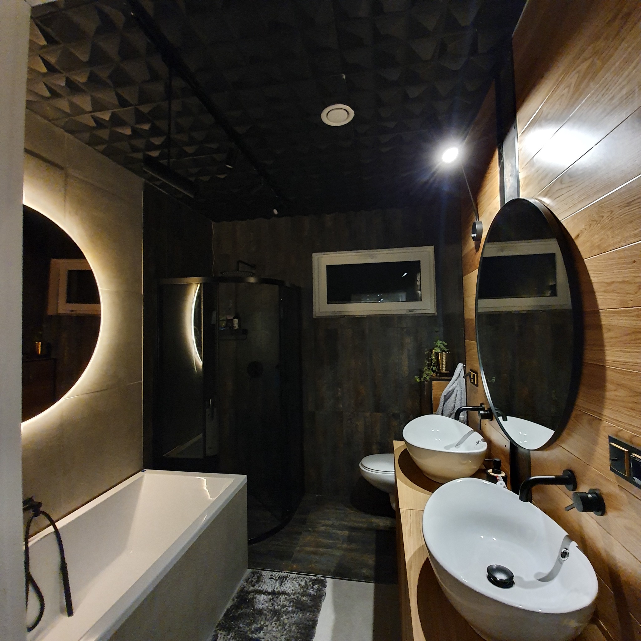 Nowoczesna łazienka z czarną kabiną prysznicową, dwiema owalnymi umywalkami nablatowymi, drewnianą okładziną ściany, lustrem z podświetleniem LED i geometrycznym wzorem na suficie.