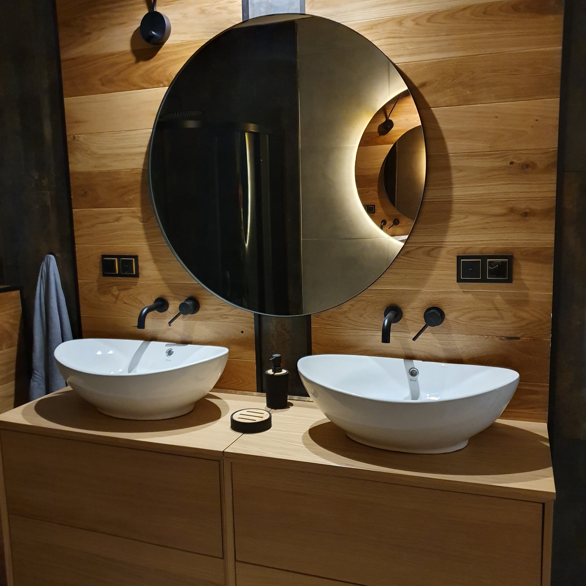 Nowoczesna łazienka z dwoma owalnymi umywalkami nablatowymi, czarną armaturą, okrągłym lustrem z podświetleniem LED i drewnianą okładziną ścienną.