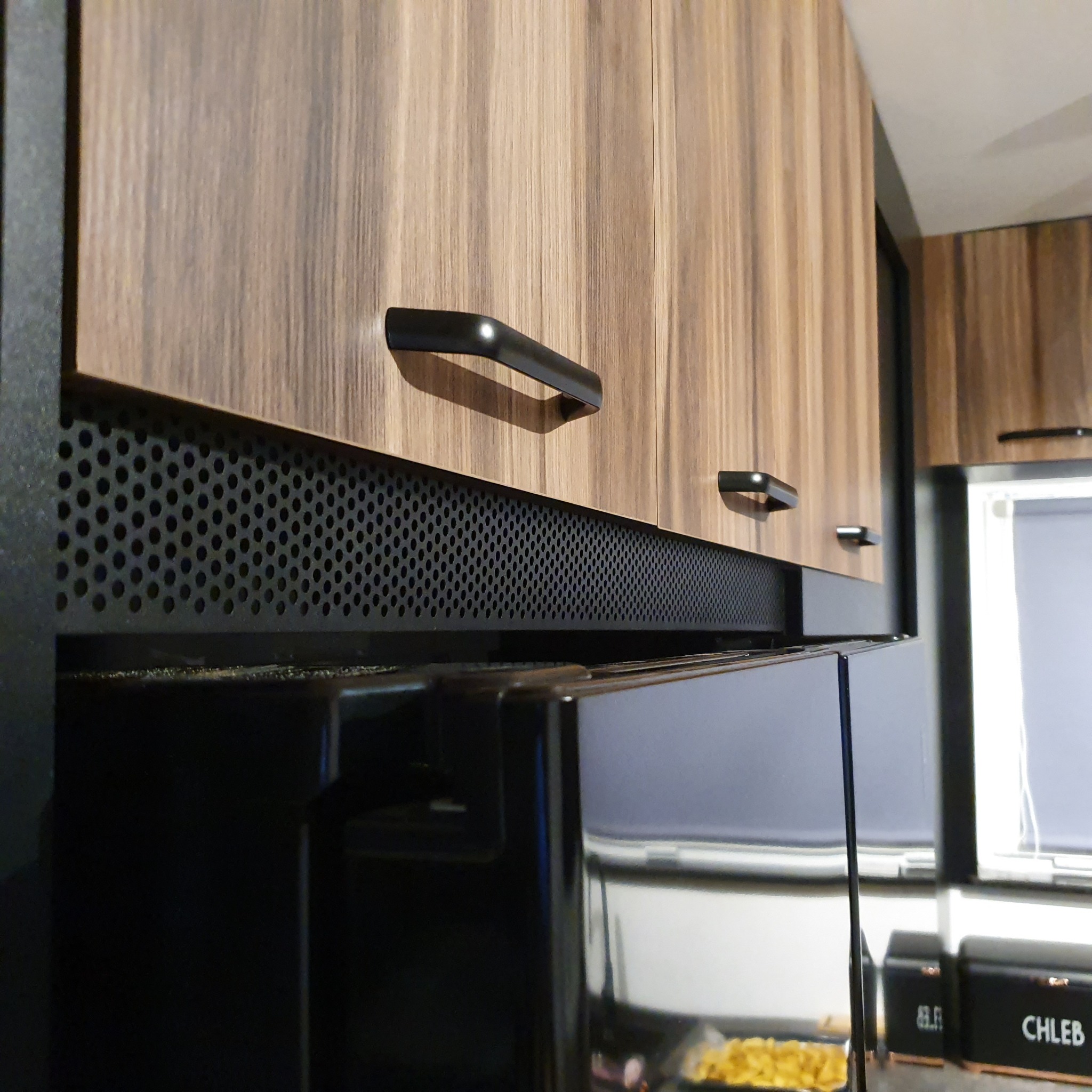 Szczegół ujęcia górnych szafek kuchennych z drewnianymi frontami i czarnymi uchwytami, z perforowanym panelem wentylacyjnym pod spodem.