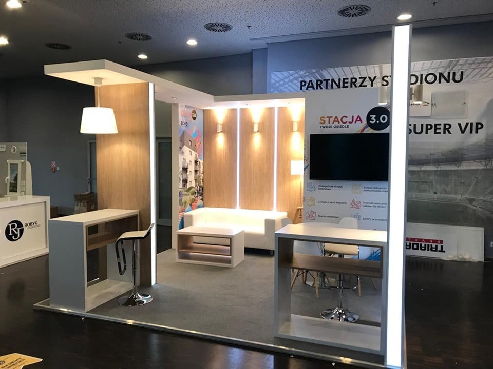 Aranżacja stoiska targowego z drewnianymi panelami, białymi meblami i podświetleniem LED, prezentująca ofertę osiedla mieszkaniowego na tle grafiki z partnerami i wizualizacją stadionu.