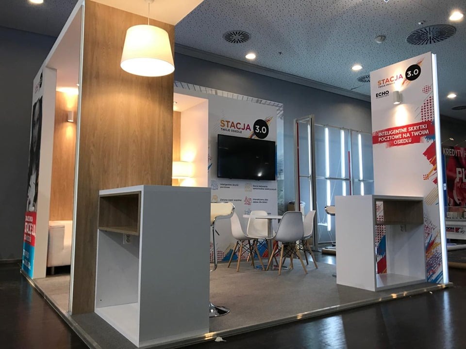 Nowoczesne stoisko targowe z elementami drewna, białymi meblami i oświetleniem punktowym, prezentujące ofertę 'Stacja 3.0'.