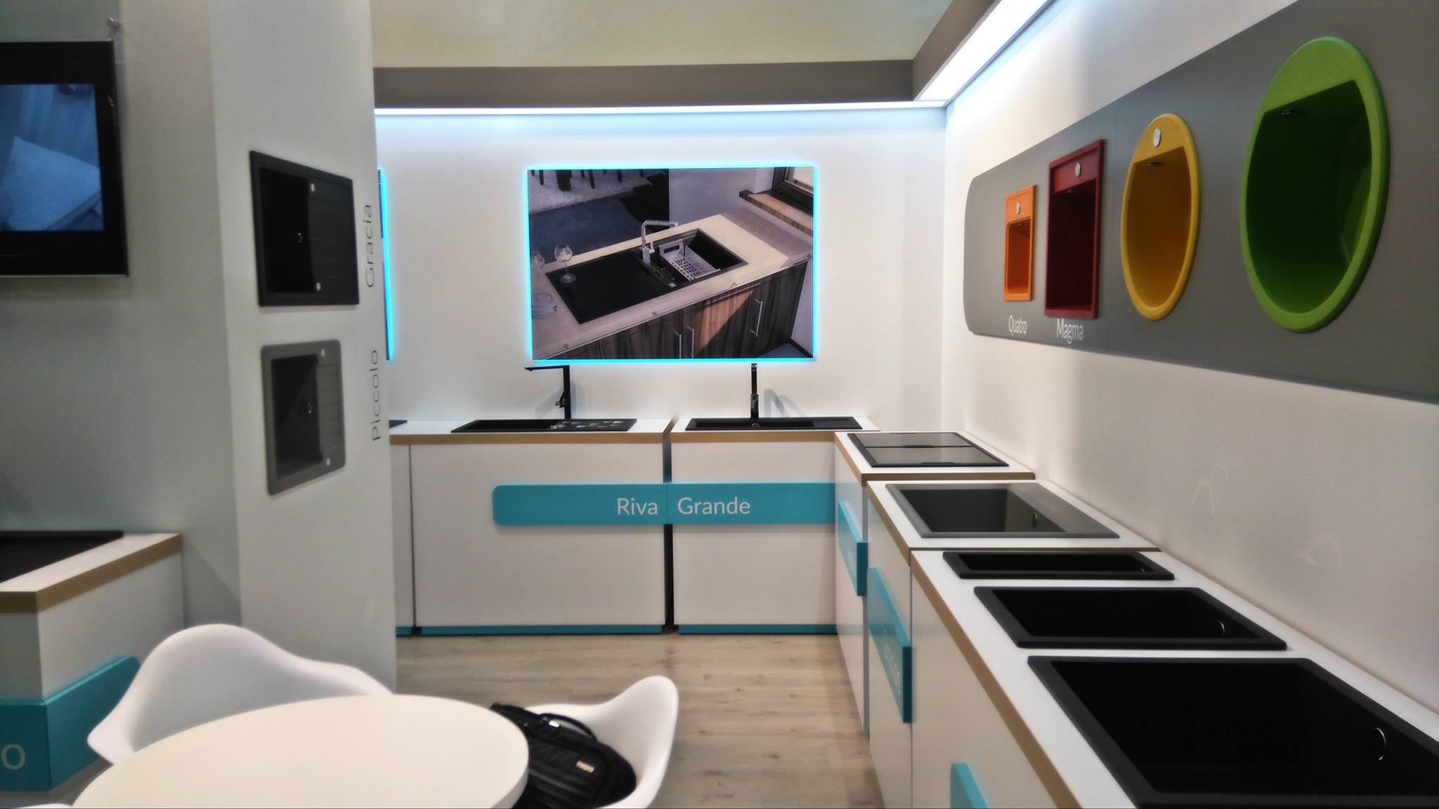 Ekspozycja zlewozmywaków kuchennych różnych modeli i kolorów w showroomie, z wyświetlaczem prezentującym aranżację kuchni z jednym z modeli, oraz oznaczeniami nazw modeli: Riva Grande, Gracia...