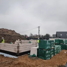 Wznoszenie ścian fundamentowych z bloczków silikatowych H+H na izolacji przeciwwilgociowej, widoczny pracownik w kamizelce odblaskowej i sterty zapakowanych bloczków na placu budowy, w tle ziemia...