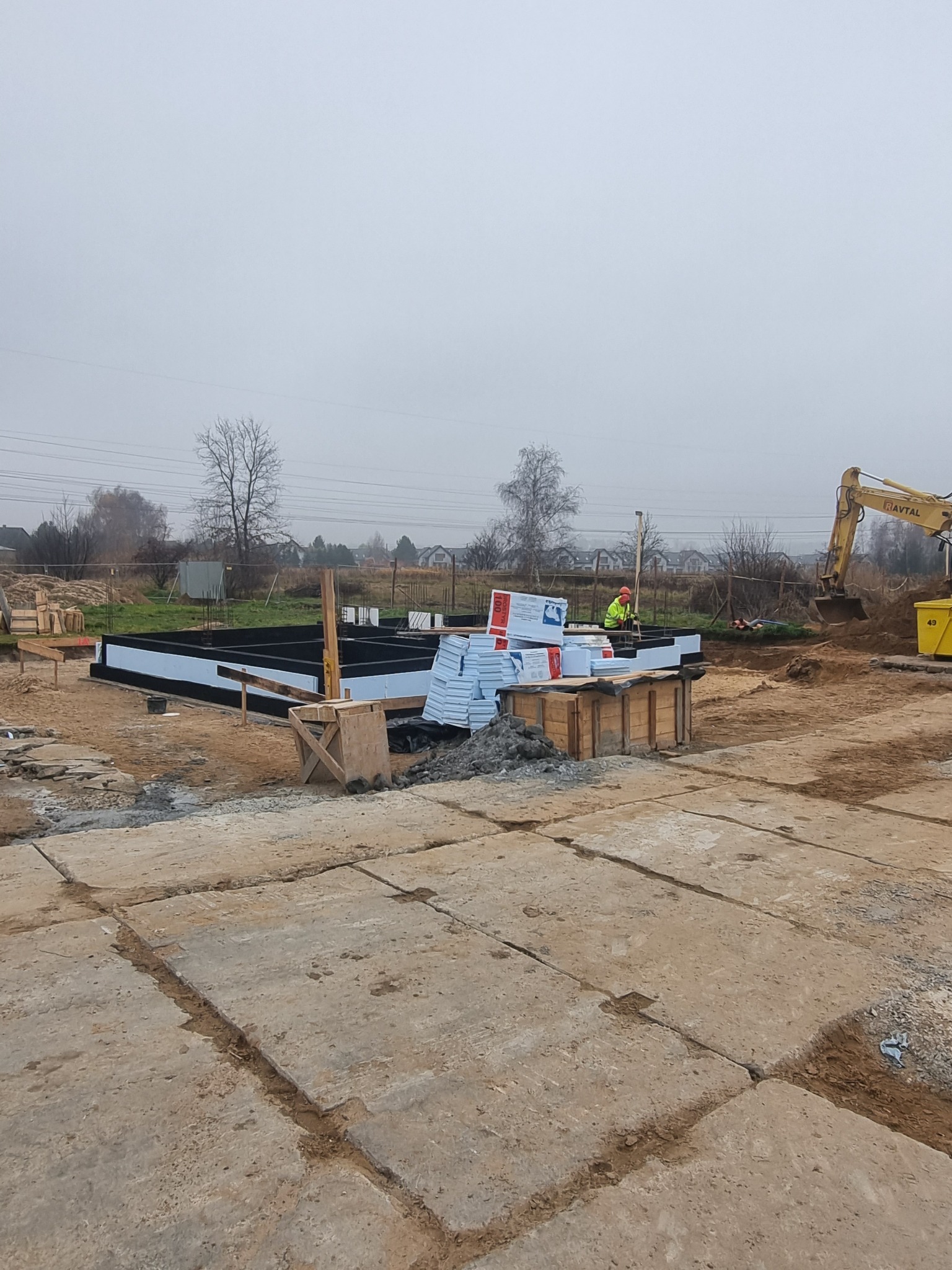 Fundamenty domu jednorodzinnego z izolacją termiczną, widoczny pracownik w odblaskowej kamizelce i koparka w tle.