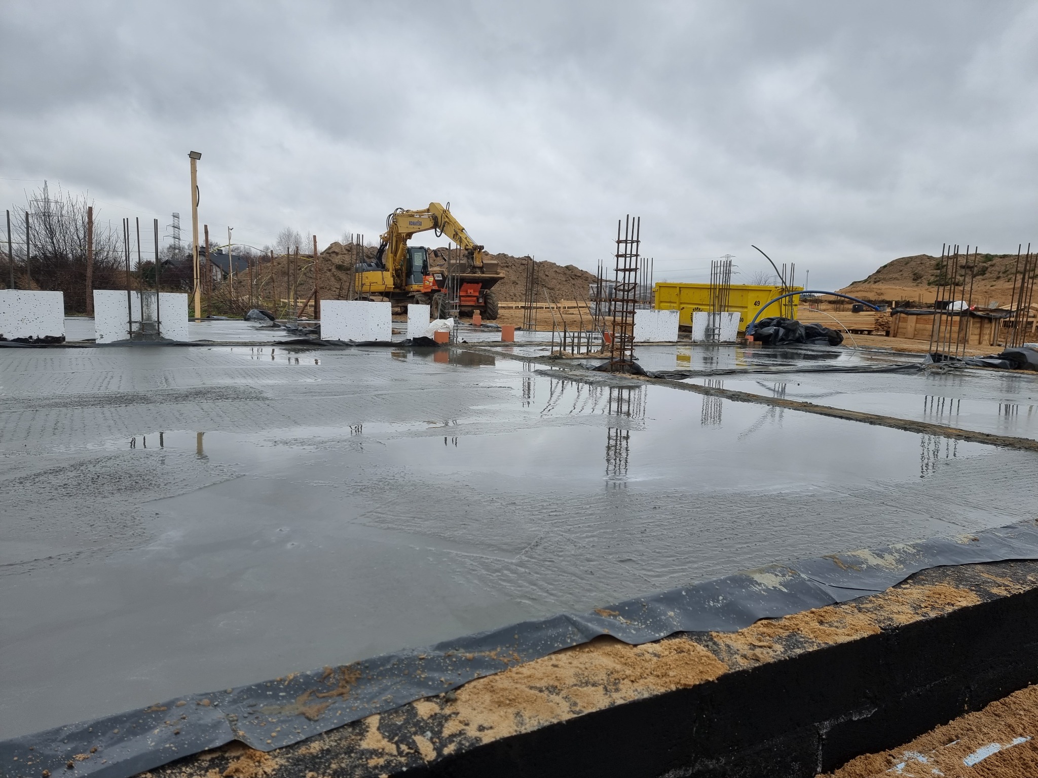 Wylany fundament pod budowę domu, zbrojenia, żółta koparka w tle, pochmurne niebo.