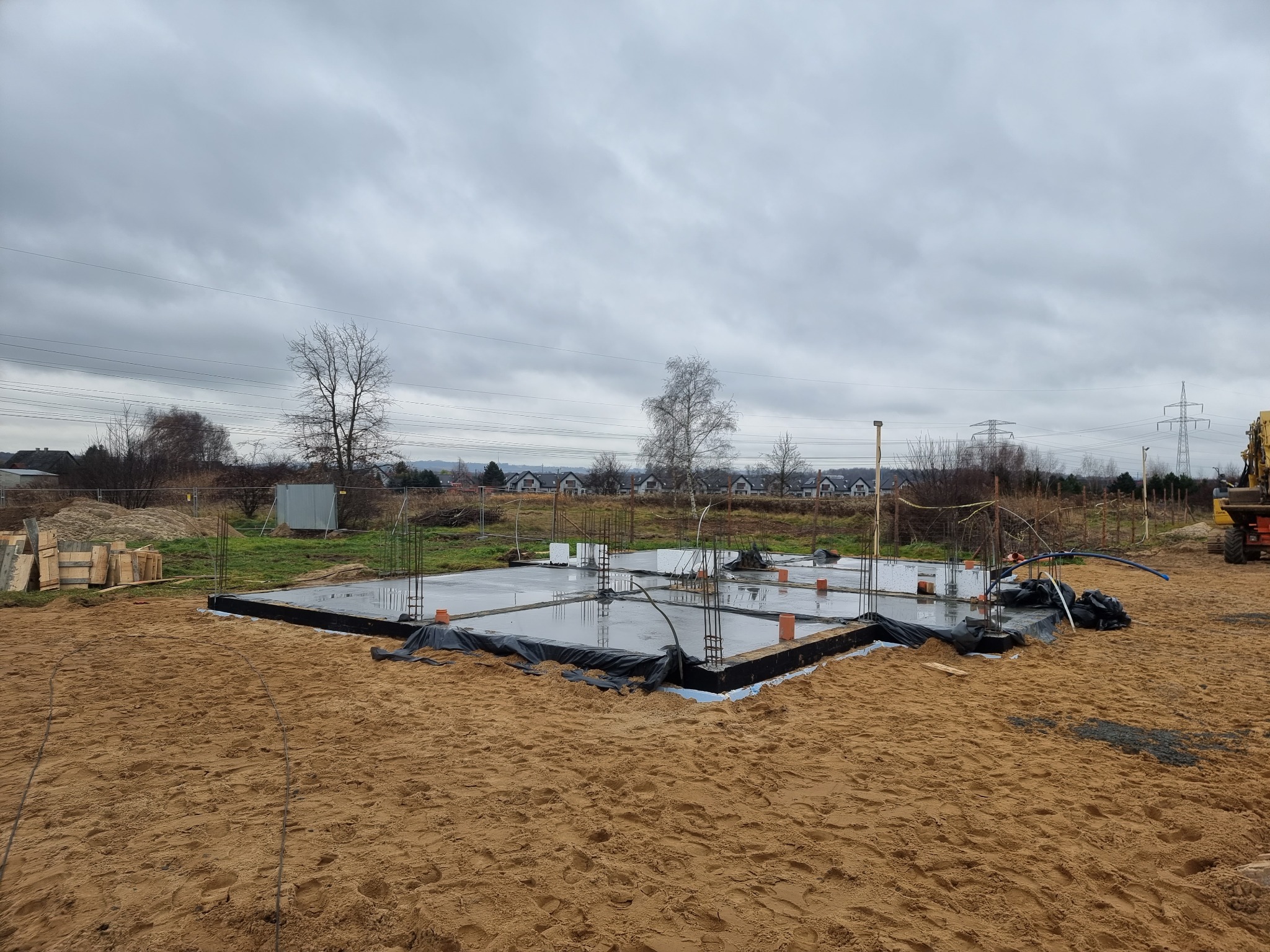Przygotowany fundament pod budowę domu: wylana płyta betonowa z wystającymi prętami zbrojeniowymi, zabezpieczona czarną folią, otoczona piaskiem, w tle pochmurne niebo i zabudowa jednorodzinna.