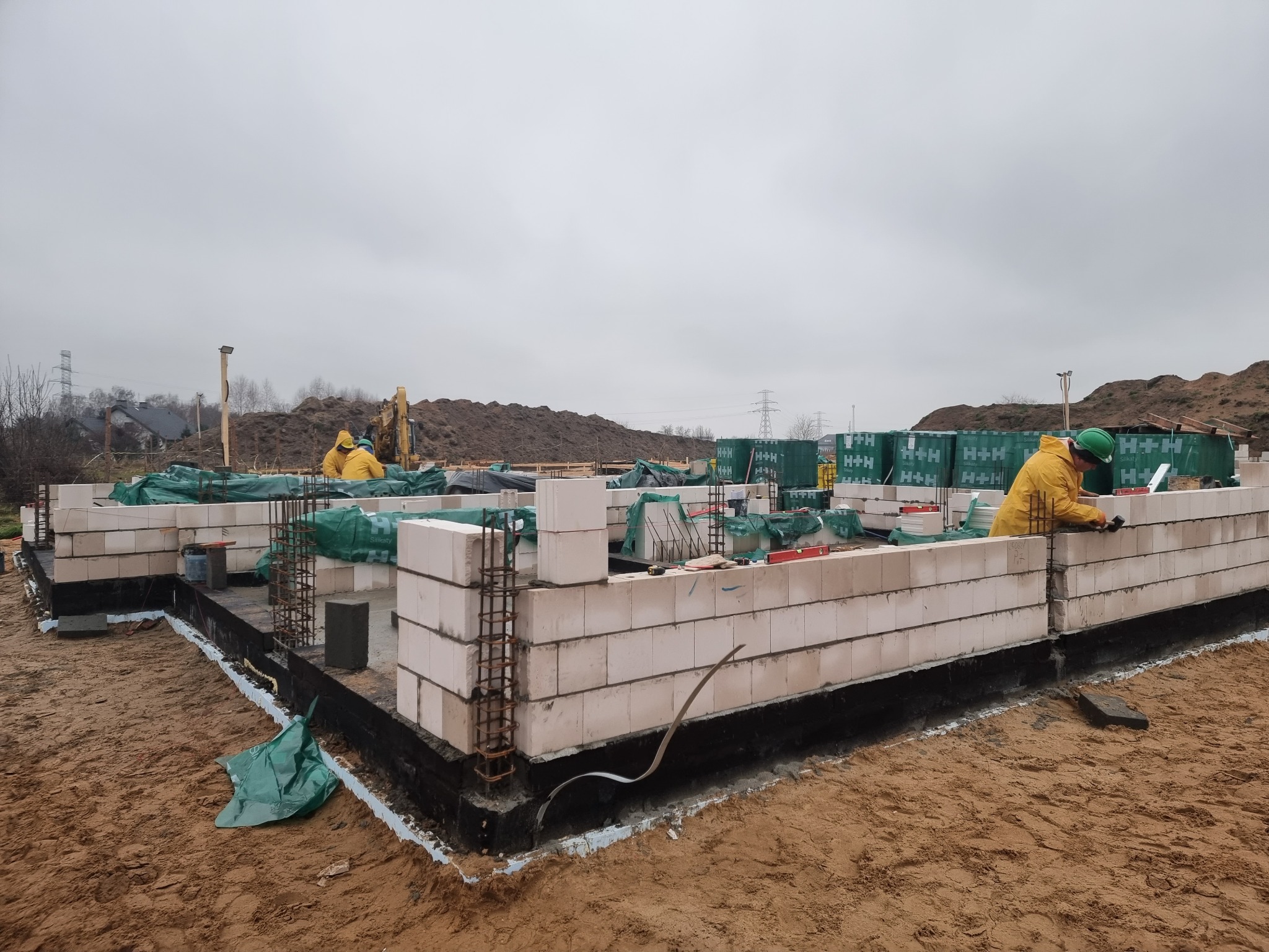 Budowa domu z bloczków silikatowych: murarz w żółtej kurtce i zielonym kasku układa pierwszą warstwę ścian na fundamencie, widoczne zbrojenia i palety bloczków H+H w tle.