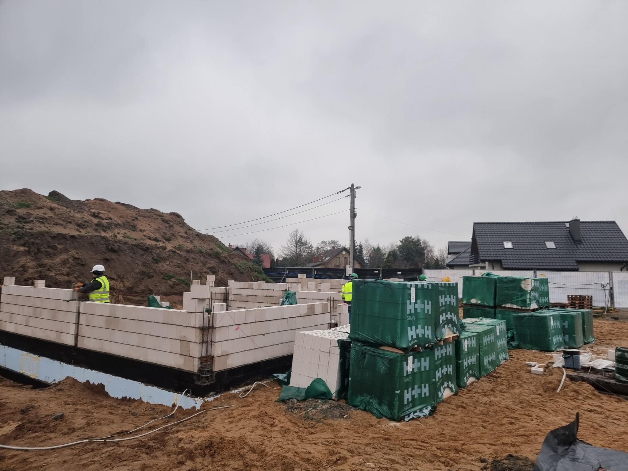 Wznoszenie ścian fundamentowych z bloczków silikatowych H+H na izolacji przeciwwilgociowej, widoczny pracownik w kamizelce odblaskowej i sterty zapakowanych bloczków na placu budowy, w tle ziemia...