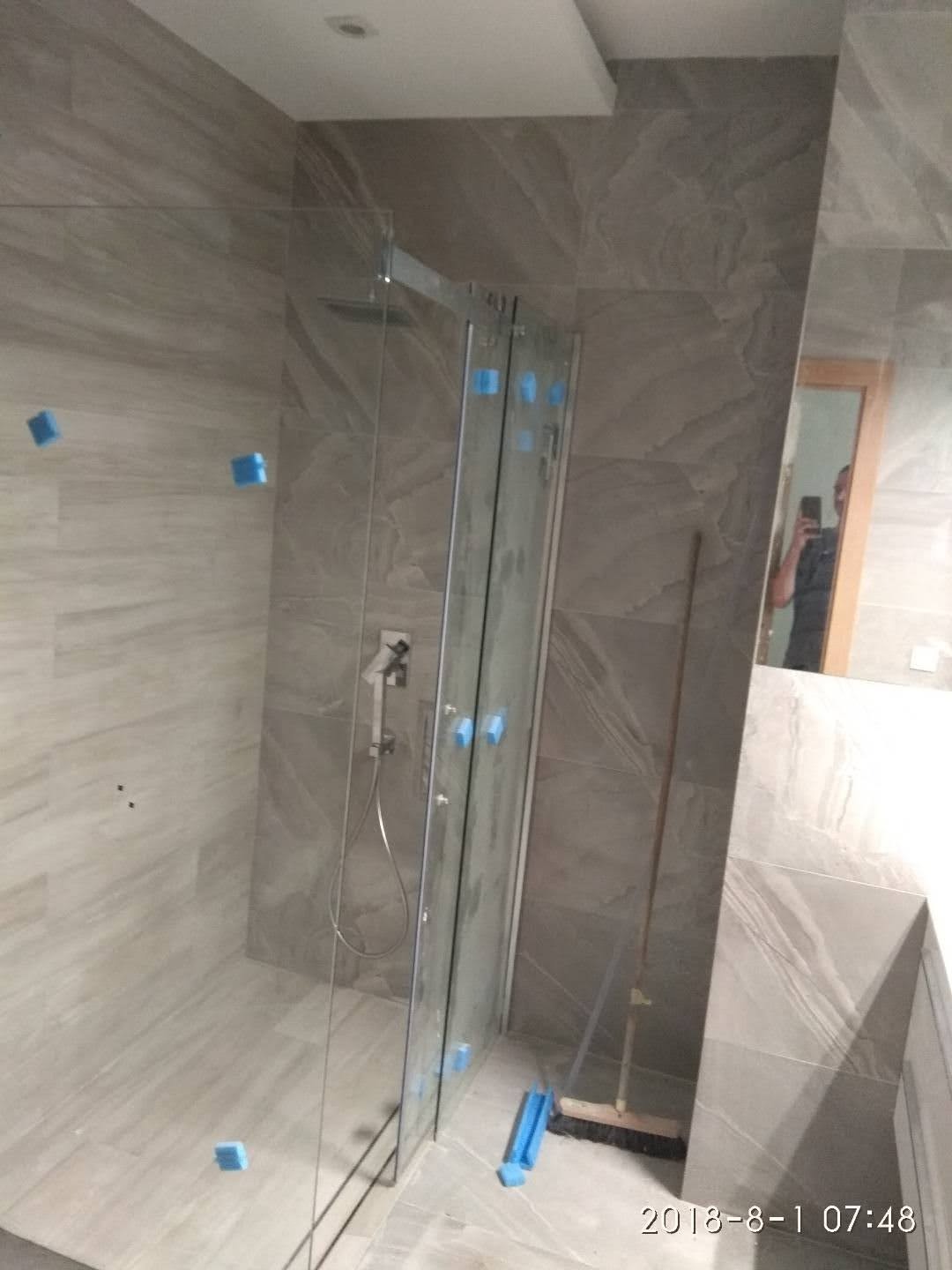 Nowoczesna kabina prysznicowa z szarymi płytkami imitującymi kamień, zabezpieczona niebieską folią, widoczna szczotka do sprzątania, odbicie osoby robiącej zdjęcie w lustrze.