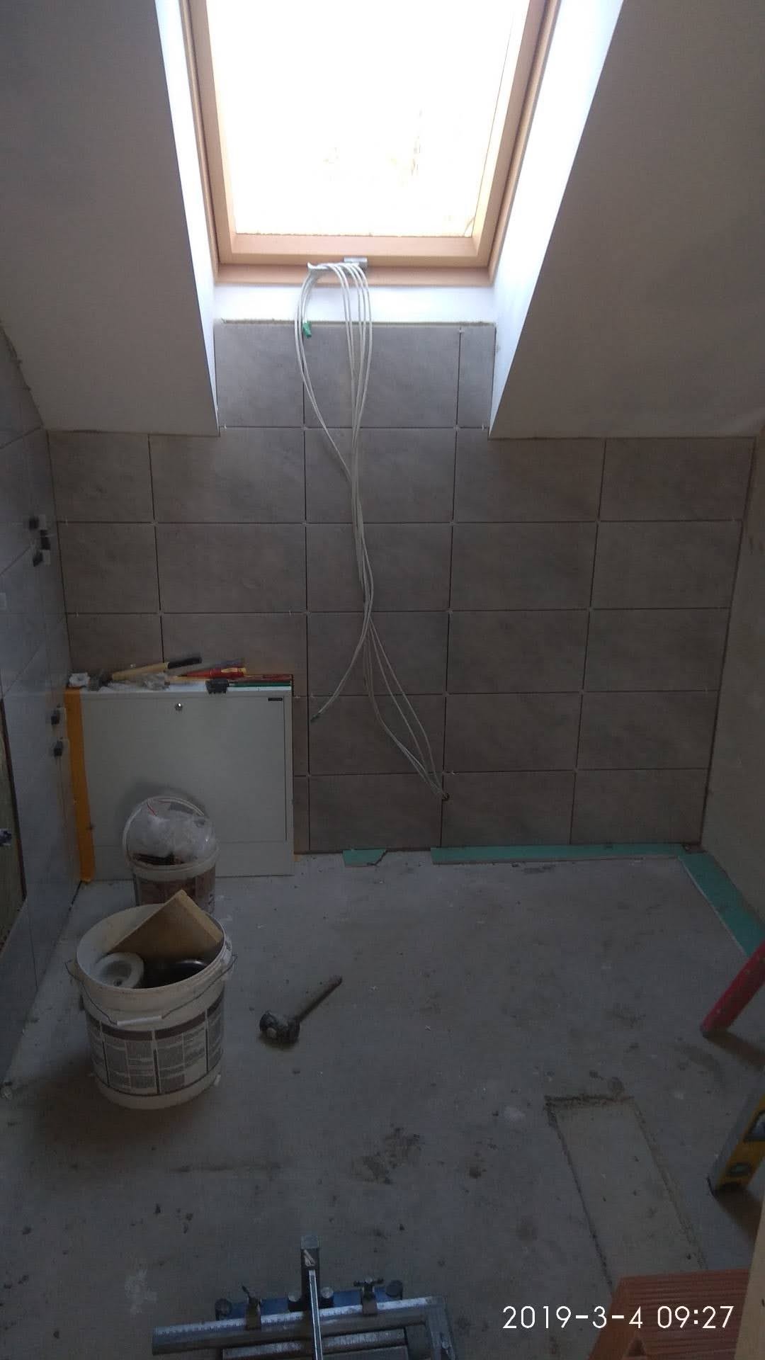 Łazienka w trakcie remontu: ułożone szare płytki na ścianach, widoczne przewody elektryczne, narzędzia i wiadro z materiałami budowlanymi na podłodze, okno dachowe w suficie.