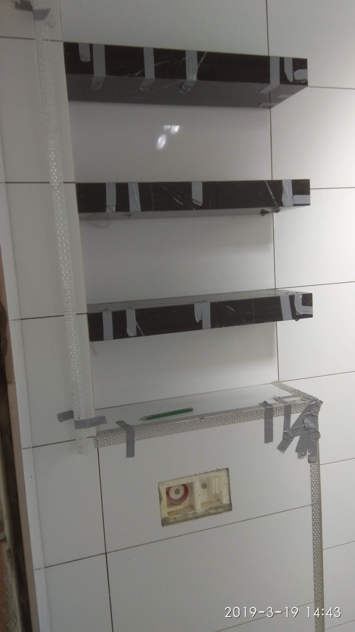 Montaż półek z czarnego marmuru w łazience, przymocowanych taśmą do białych płytek, widoczny stelaż podtynkowy WC i ołówek na metalowym profilu.