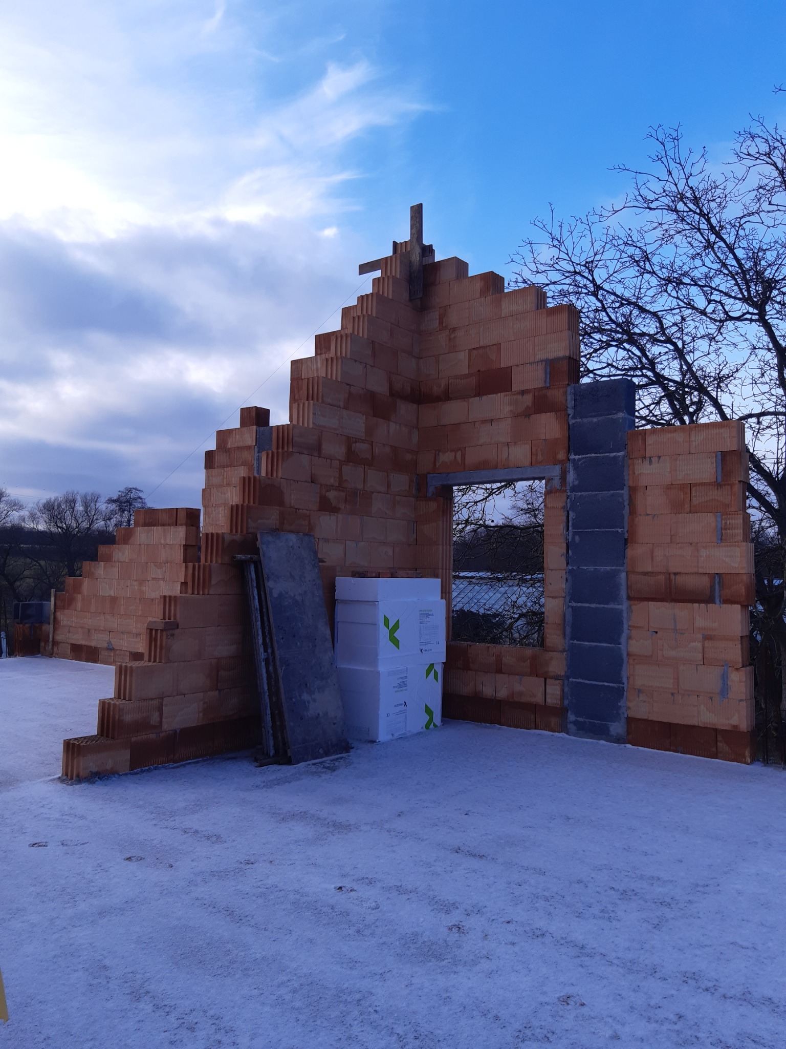 Wznoszenie ścian domu z cegły ceramicznej w zimowej scenerii, widoczne bloczki izolacyjne i szare nadproże okienne.