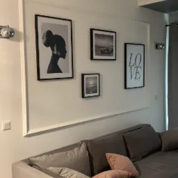 Szary narożnik z poduszkami w salonie z galerią czarno-białych obrazów w białej ramie na ścianie.