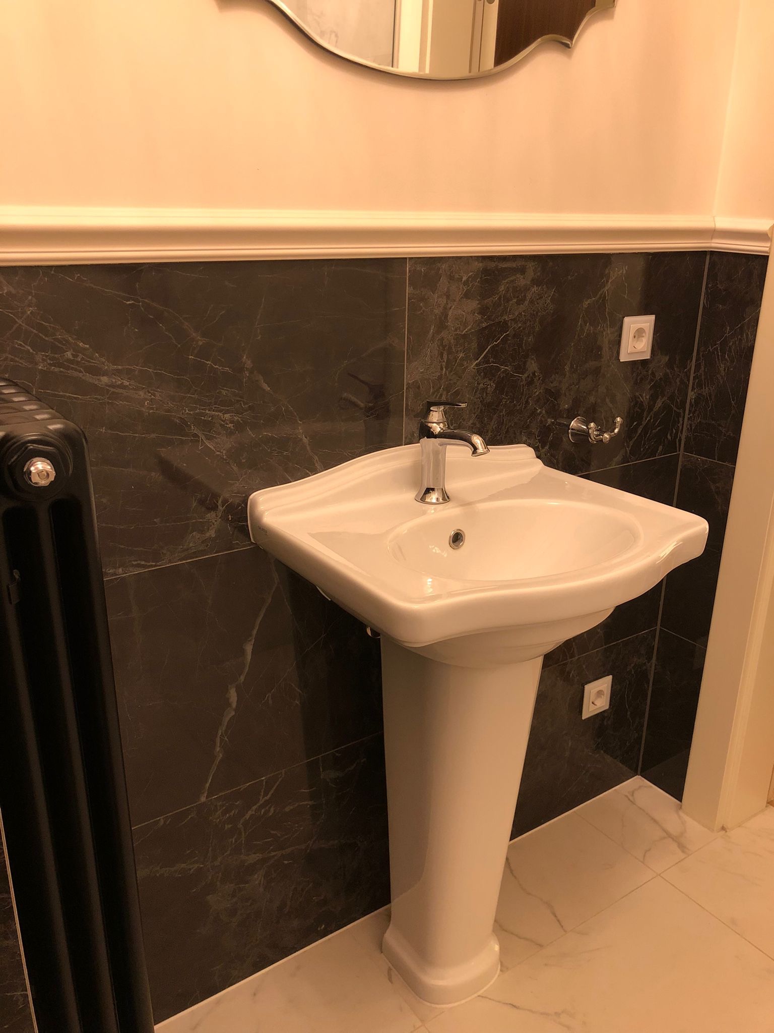 Biała umywalka na postumencie w łazience z czarnymi marmurowymi płytkami, widoczny fragment grzejnika i lustra.