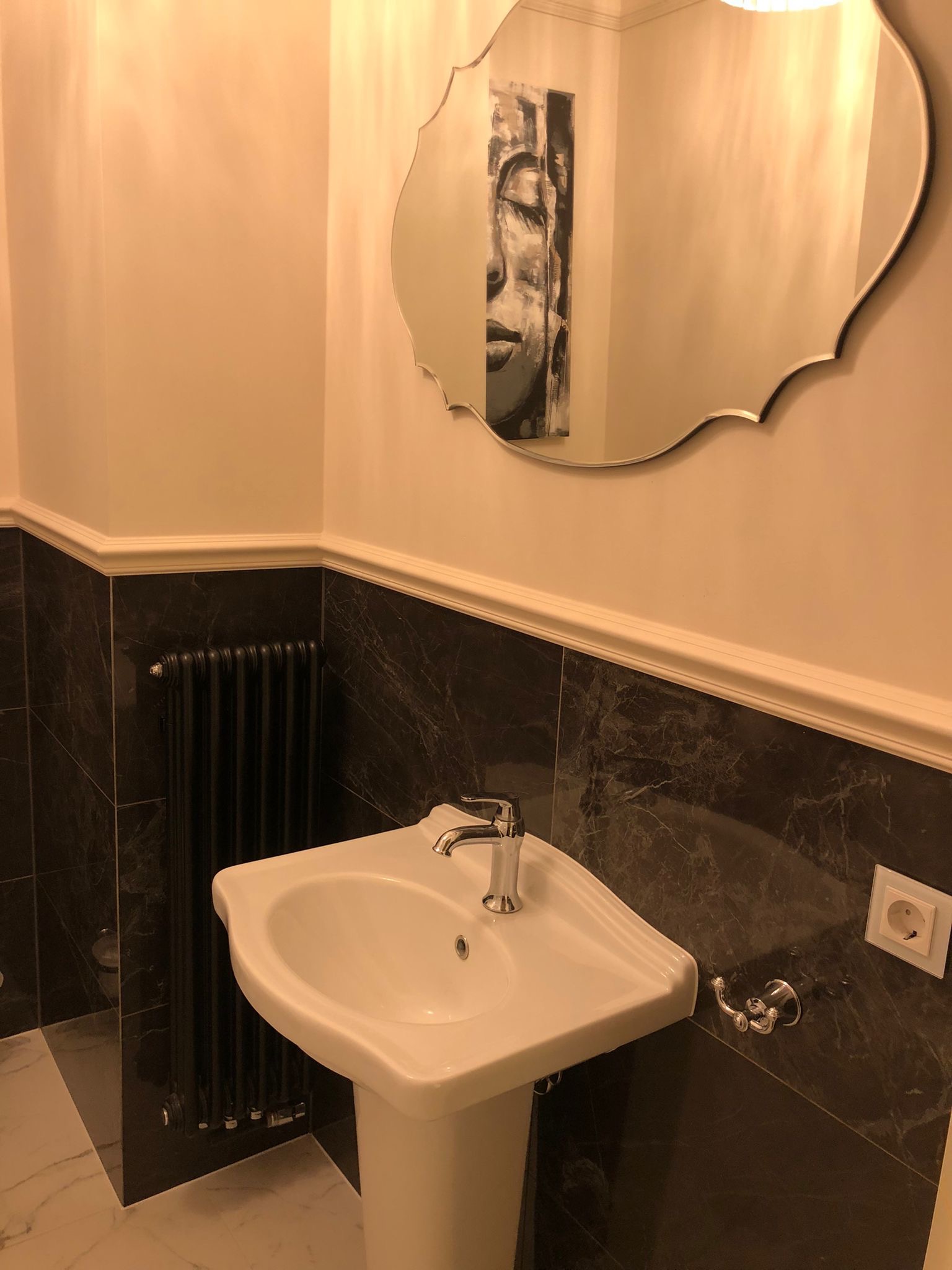 Biała umywalka na postumencie w łazience z ciemnymi marmurowymi płytkami, czarnym grzejnikiem żeberkowym i ozdobnym lustrem odbijającym obraz