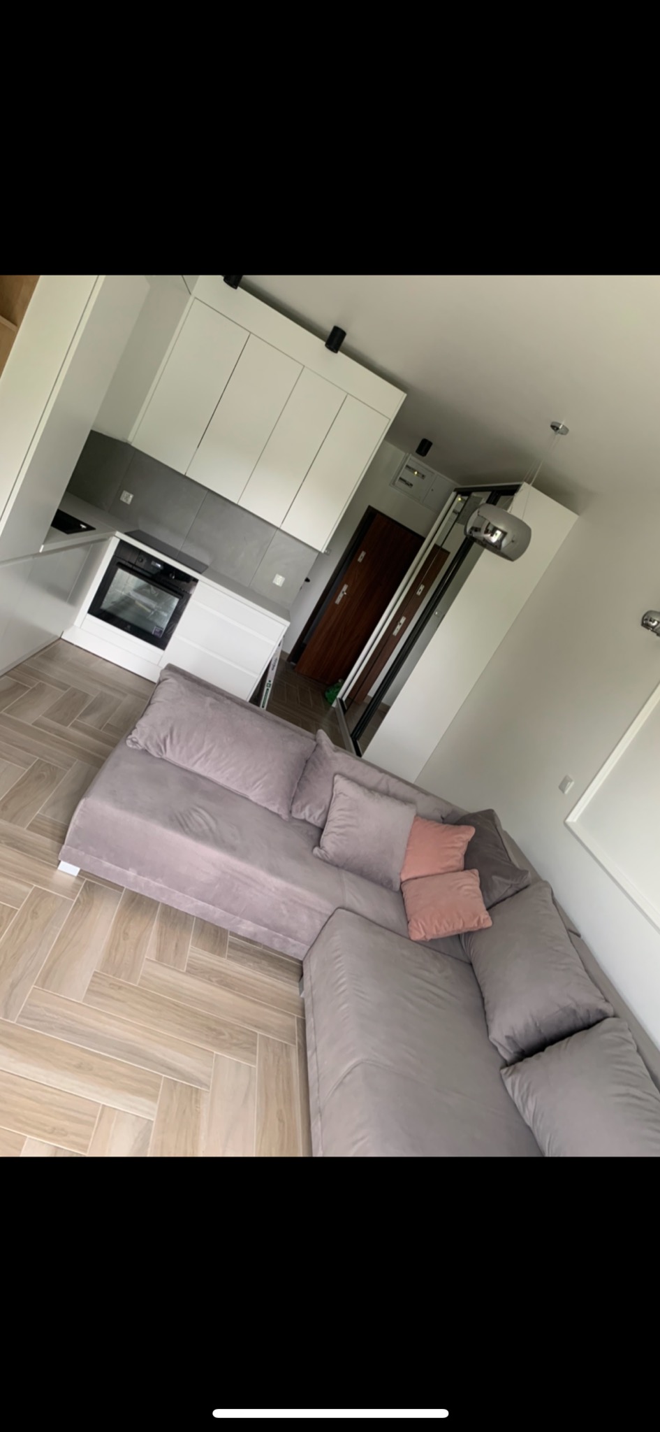 Nowoczesna kuchnia z białymi szafkami, szarymi płytkami i jasną podłogą w jodełkę, widoczna z perspektywy narożnika szarej sofy z poduszkami.