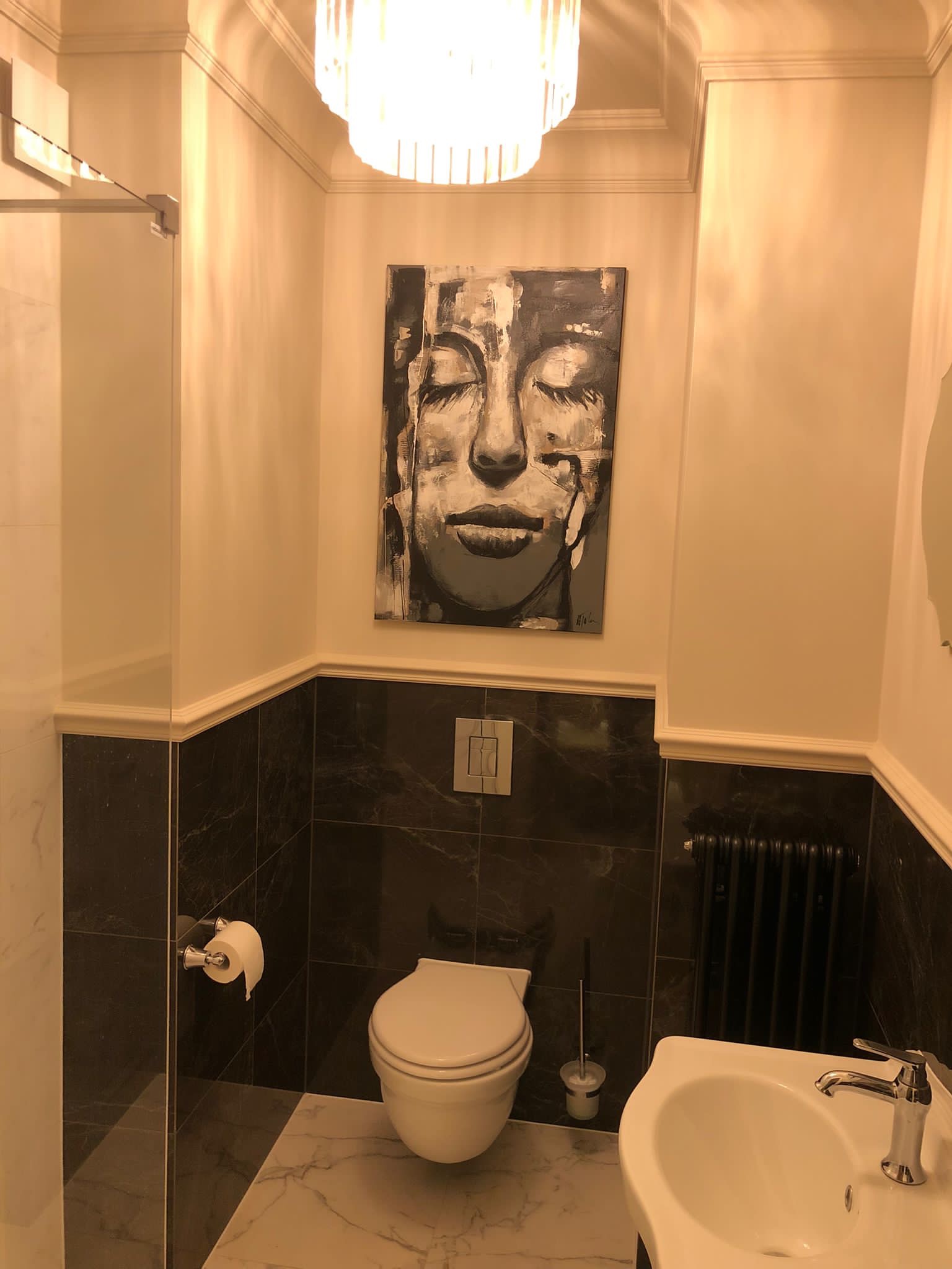 Ukończona łazienka z czarnymi marmurowymi płytkami do połowy ściany, białą ceramiką, grzejnikiem żeberkowym i obrazem kobiety nad toaletą pod ozdobnym żyrandolem.