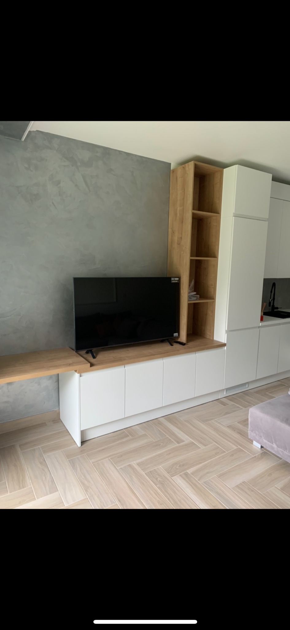 Nowoczesny salon z szarą ścianą z efektem dekoracyjnym, białą zabudową meblową z drewnianymi akcentami, telewizorem i podłogą w jodełkę.