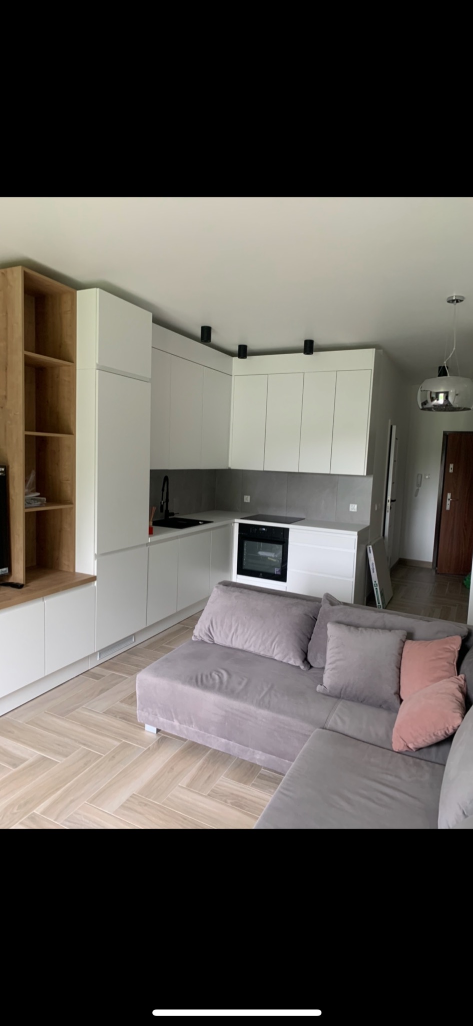 Nowoczesna kuchnia z białymi szafkami, szarym blatem i czarną armaturą, widok z salonu z szarą sofą i parkietem ułożonym w jodełkę.