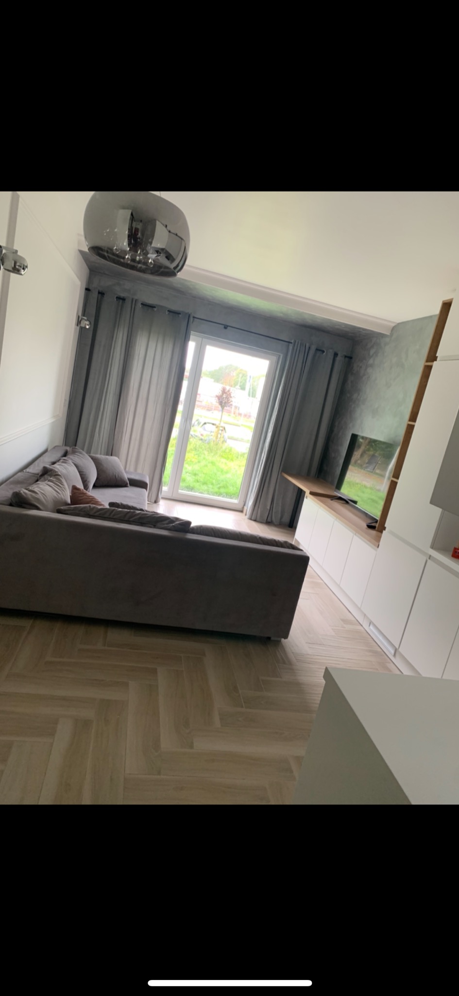 Nowoczesny salon z szarą sofą narożną, parkietem ułożonym w jodełkę, szarymi zasłonami i błyszczącą, okrągłą lampą sufitową