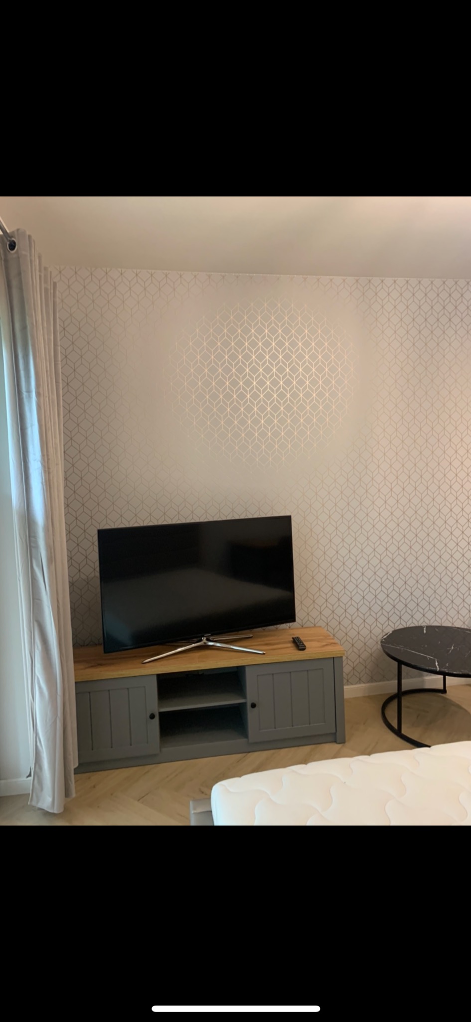 Pokój dzienny z telewizorem na szarej szafce RTV, wzorzysta tapeta w geometryczne kształty, stolik kawowy z czarnym marmurowym blatem i fragment łóżka z białym materacem.