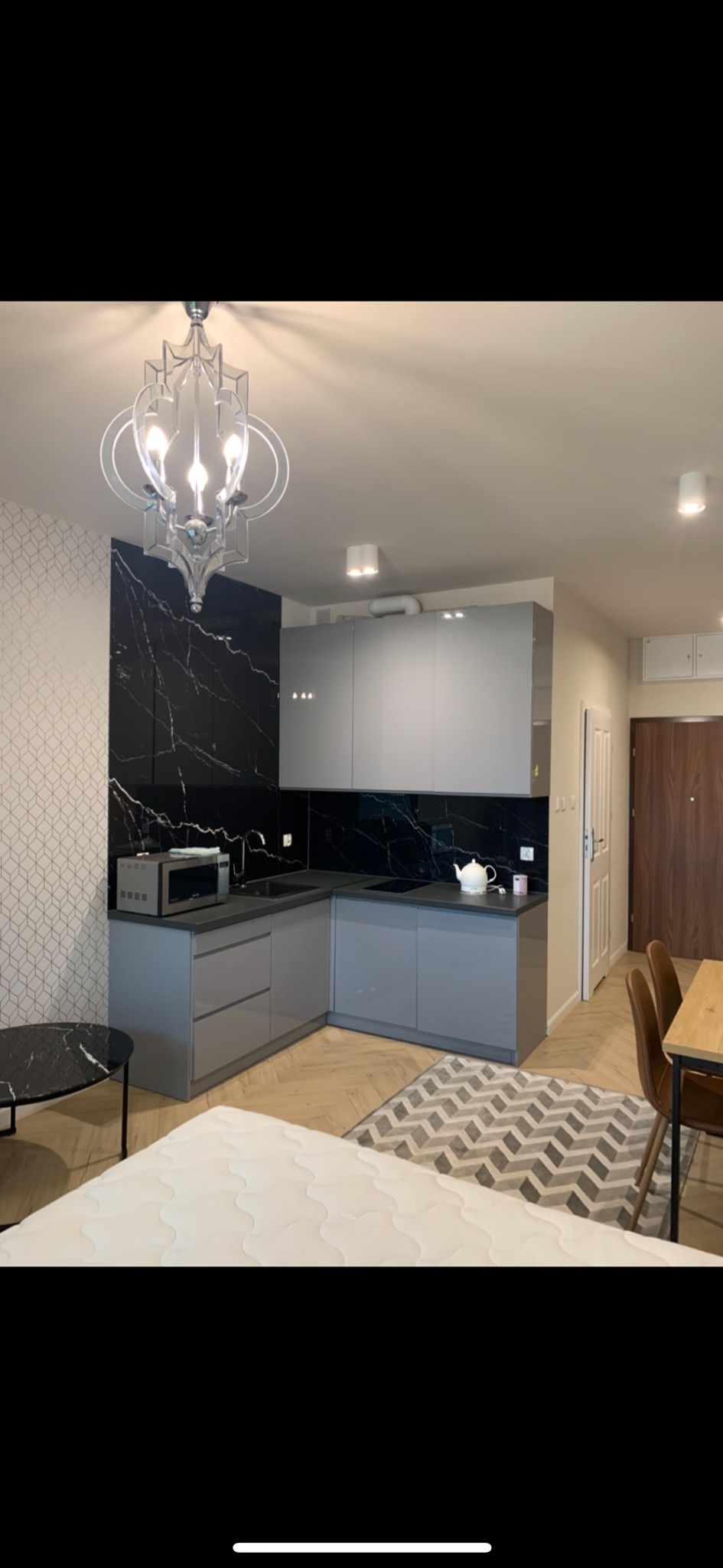 Szara kuchnia w aneksie, czarny marmur na ścianie, ozdobny żyrandol, widok z perspektywy łóżka, dywan w zygzaki na parkiecie.