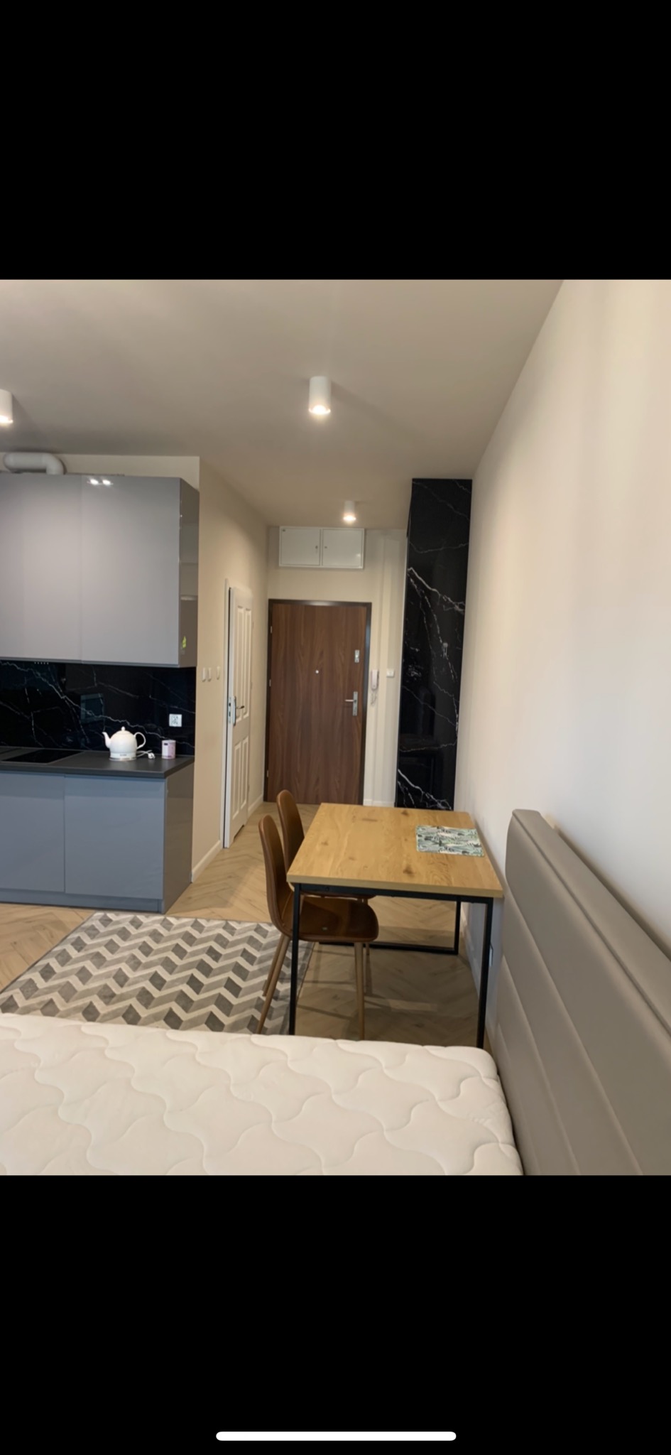 Wnętrze kawalerki z aneksem kuchennym, drewnianym stołem i krzesłami, dywanem w geometryczne wzory oraz fragmentem łóżka z pikowanym zagłówkiem.