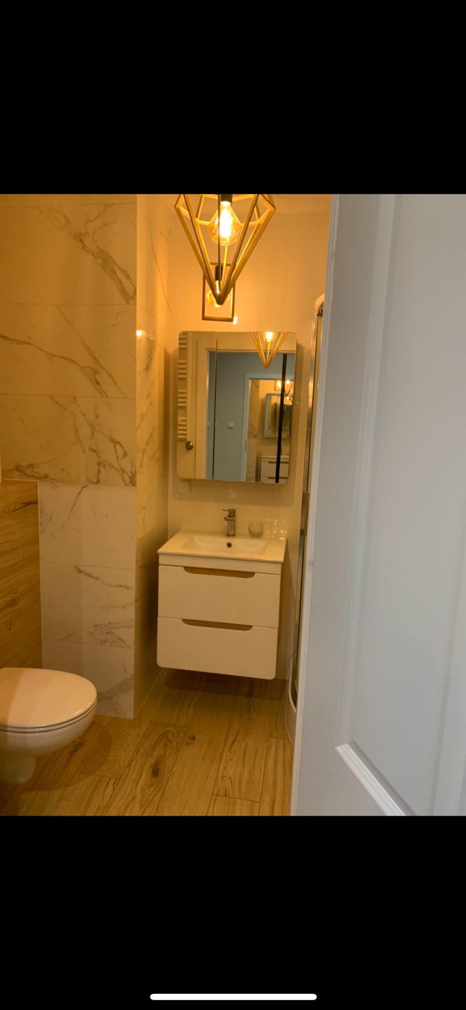 Nowoczesna łazienka z marmurowymi płytkami, drewnopodobną podłogą, wiszącą szafką z umywalką, lustrem z oświetleniem i dekoracyjną lampą wiszącą w kształcie diamentu.